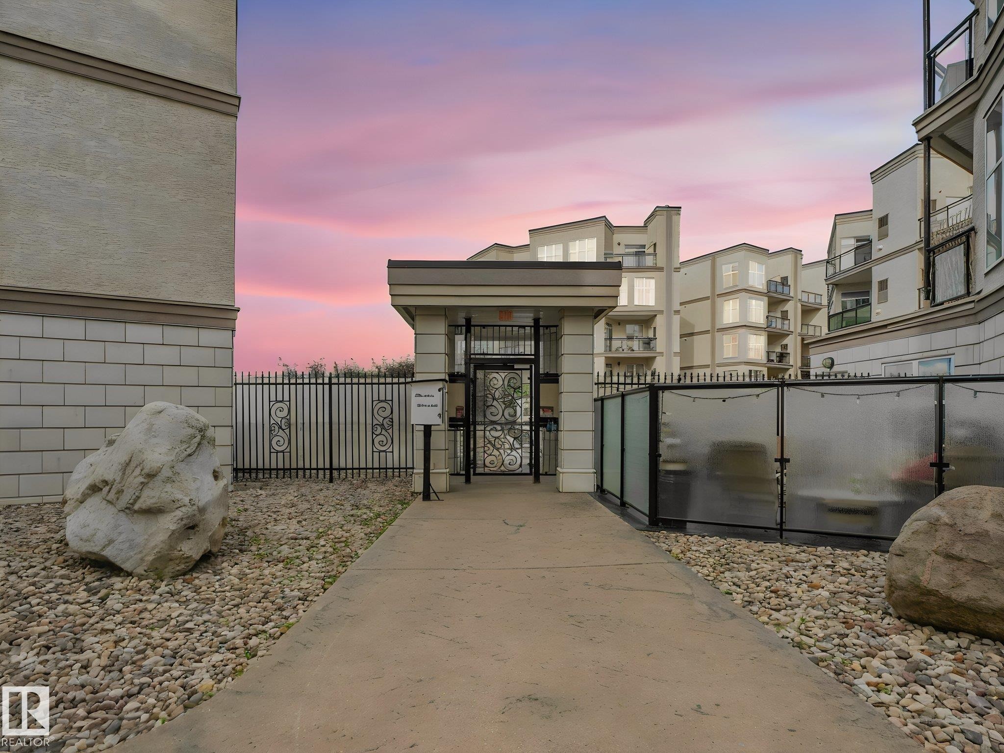 Gated entry featuring decorative wrought ironwork, concrete pathway, and surrounding gravel landscaping with large boulders - 150 4827 104A Street, Edmonton, AB