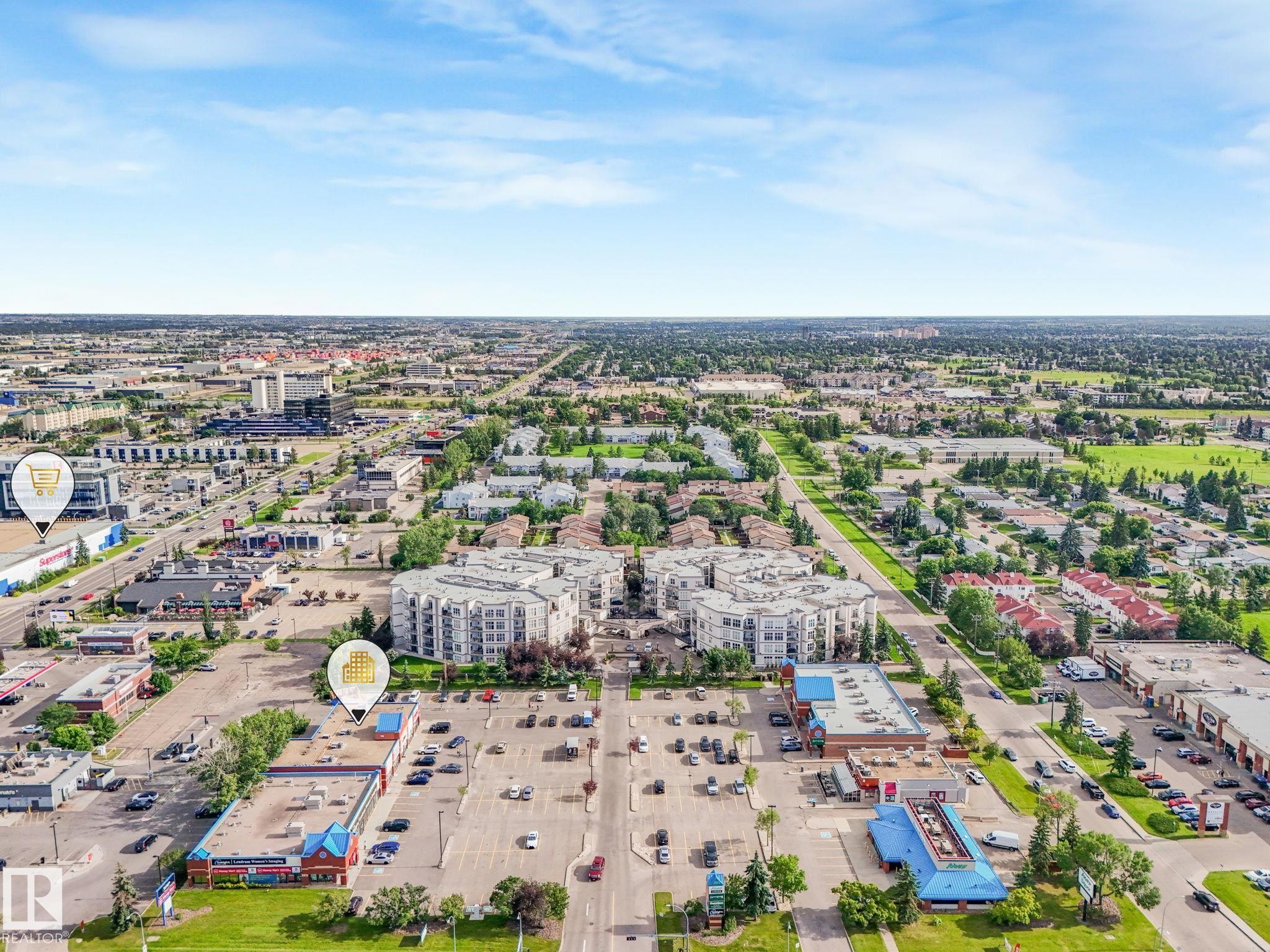 Aerial perspective showcasing a mixed-use urban landscape featuring commercial structures, multi-story residential buildings, and extensive paved parking areas - 150 4827 104A Street, Edmonton, AB