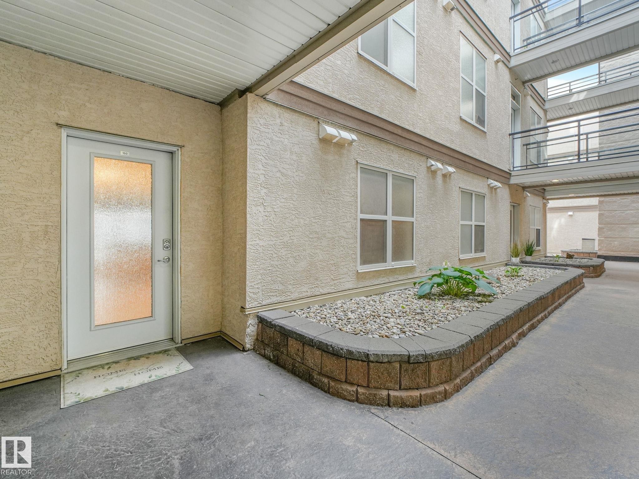 Ground-level entry featuring a frosted glass door, textured stucco facade, and a decorative concrete pathway - 150 4827 104A Street, Edmonton, AB