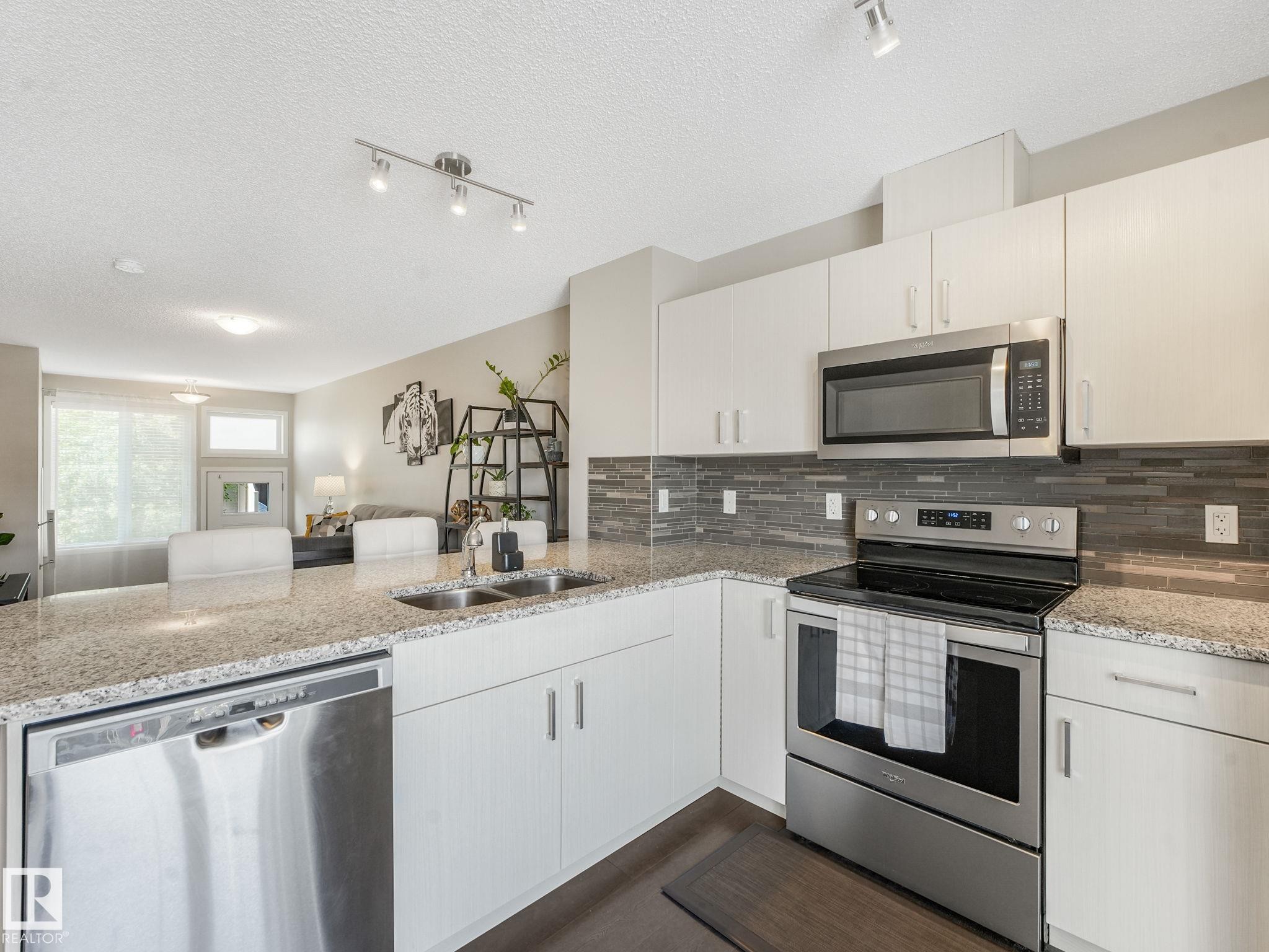 Modern kitchen featuring light-toned cabinetry, granite-finish countertops, stainless steel appliances, a grey subway tile backsplash, and a double basin sink - #55 4470 Prowse Rd Sw, Edmonton, AB - Indoor Photo Showing Kitchen With Stainless Steel Kitchen With Double Sink With Upgraded Kitchen