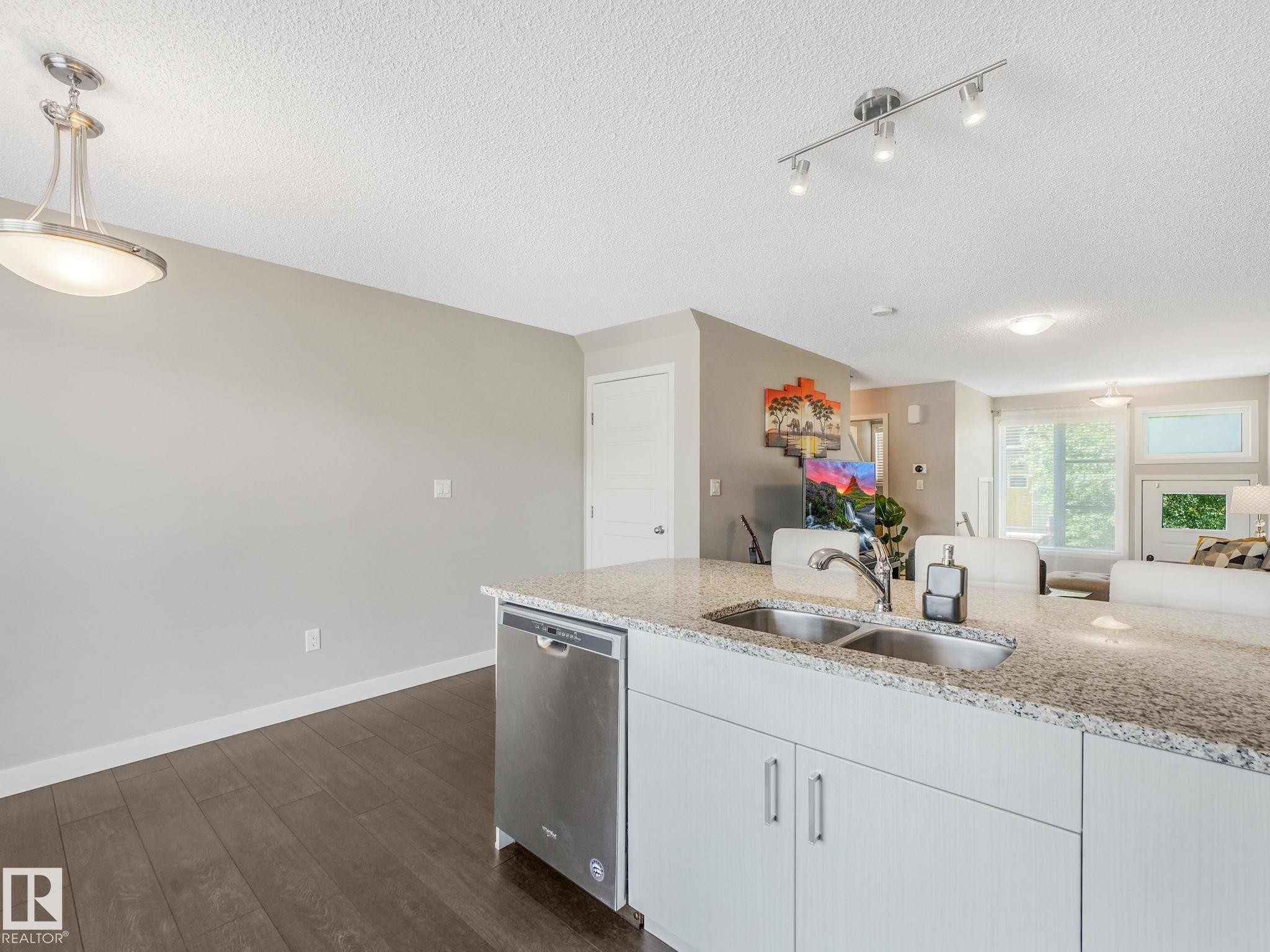 Kitchen island featuring granite countertops, a double-basin stainless steel sink with a high-arc faucet, and integrated stainless steel dishwasher - #55 4470 Prowse Rd Sw, Edmonton, AB - Indoor Photo Showing Kitchen With Double Sink