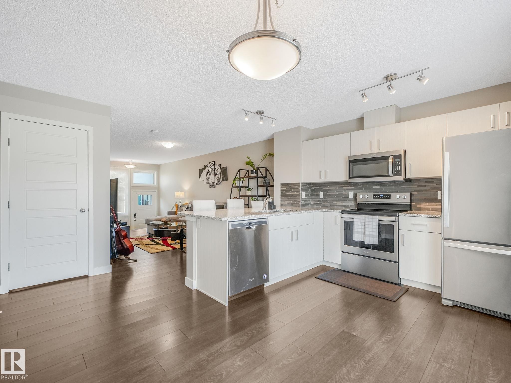 Open-concept kitchen featuring wood-finish flooring, white cabinetry, stainless steel appliances, a stone countertop, and a tiled backsplash - #55 4470 Prowse Rd Sw, Edmonton, AB - Indoor Photo Showing Kitchen With Stainless Steel Kitchen