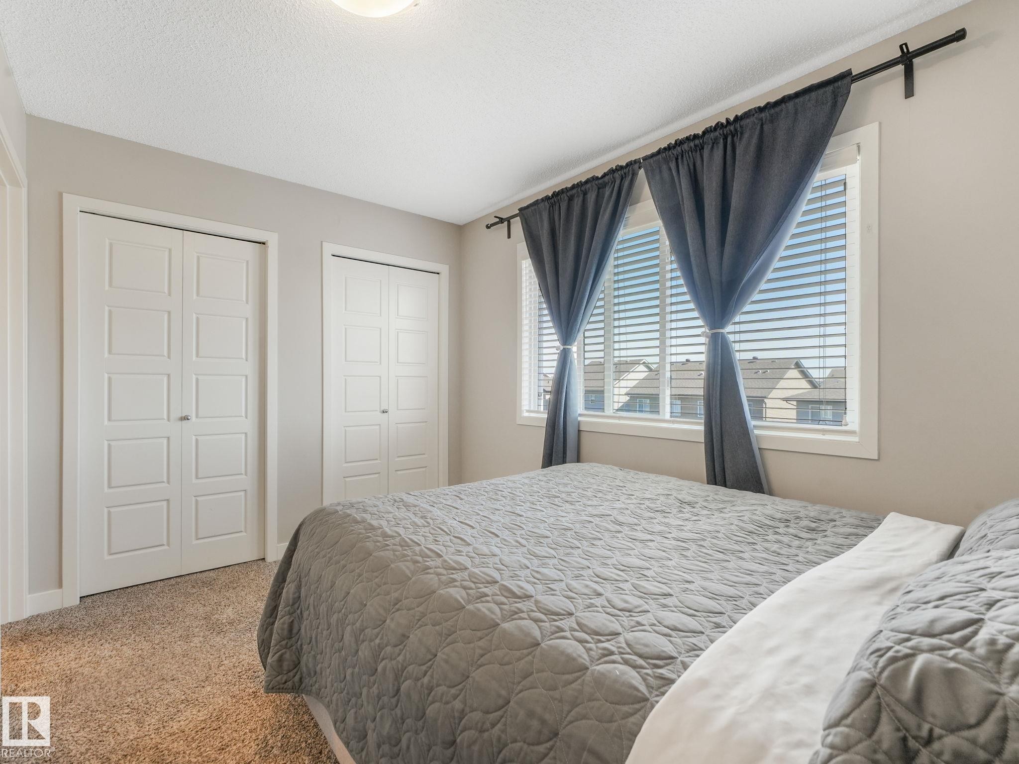 Carpeted room featuring dual bi-fold closet doors with square paneling, a large window with horizontal blinds, and a flush mount ceiling light fixture - #55 4470 Prowse Rd Sw, Edmonton, AB - Indoor Photo Showing Bedroom