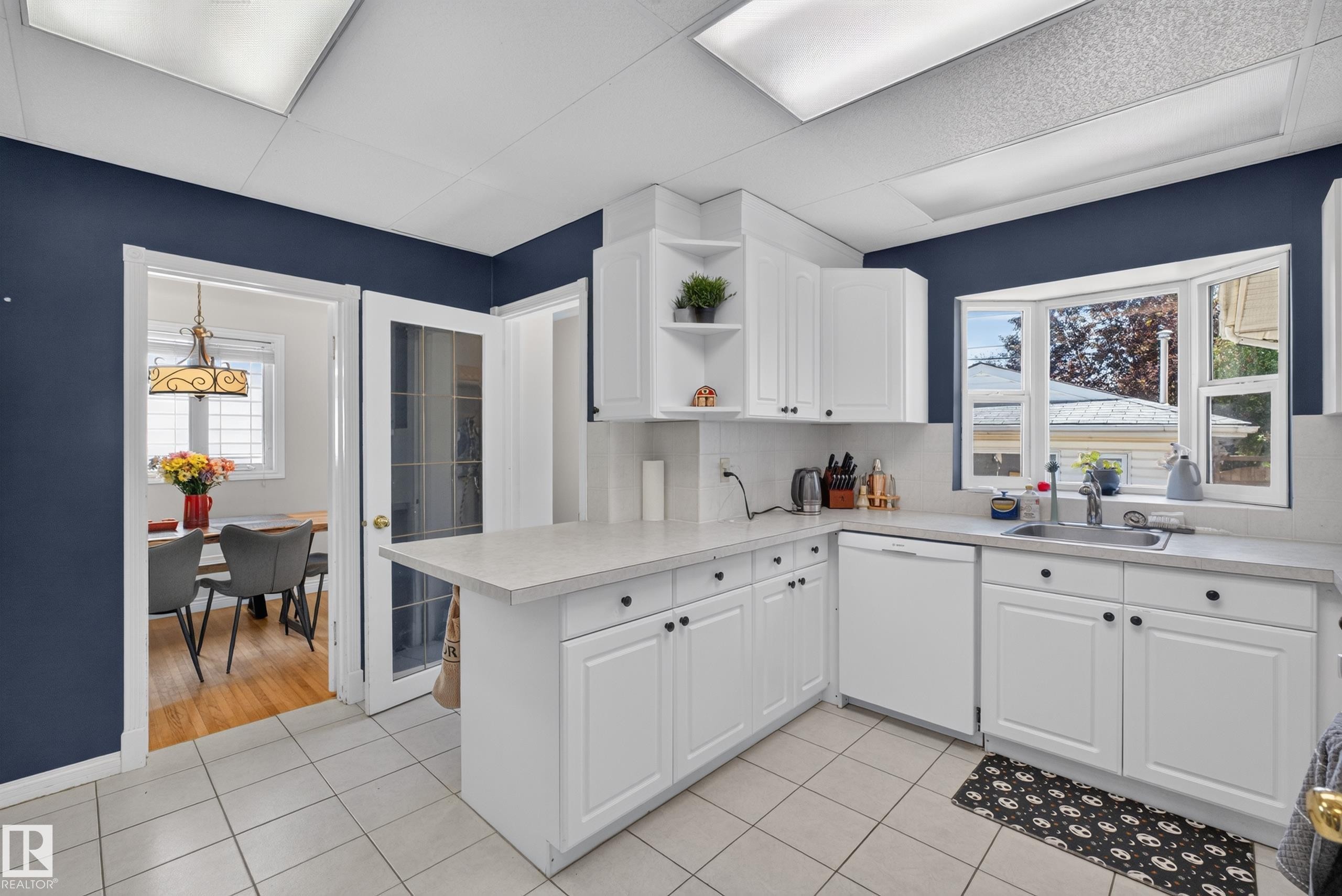 Kitchen featuring white cabinetry, light-toned countertops, a stainless steel sink, a bay window, and tile flooring - 11015 122 Street, Edmonton, AB