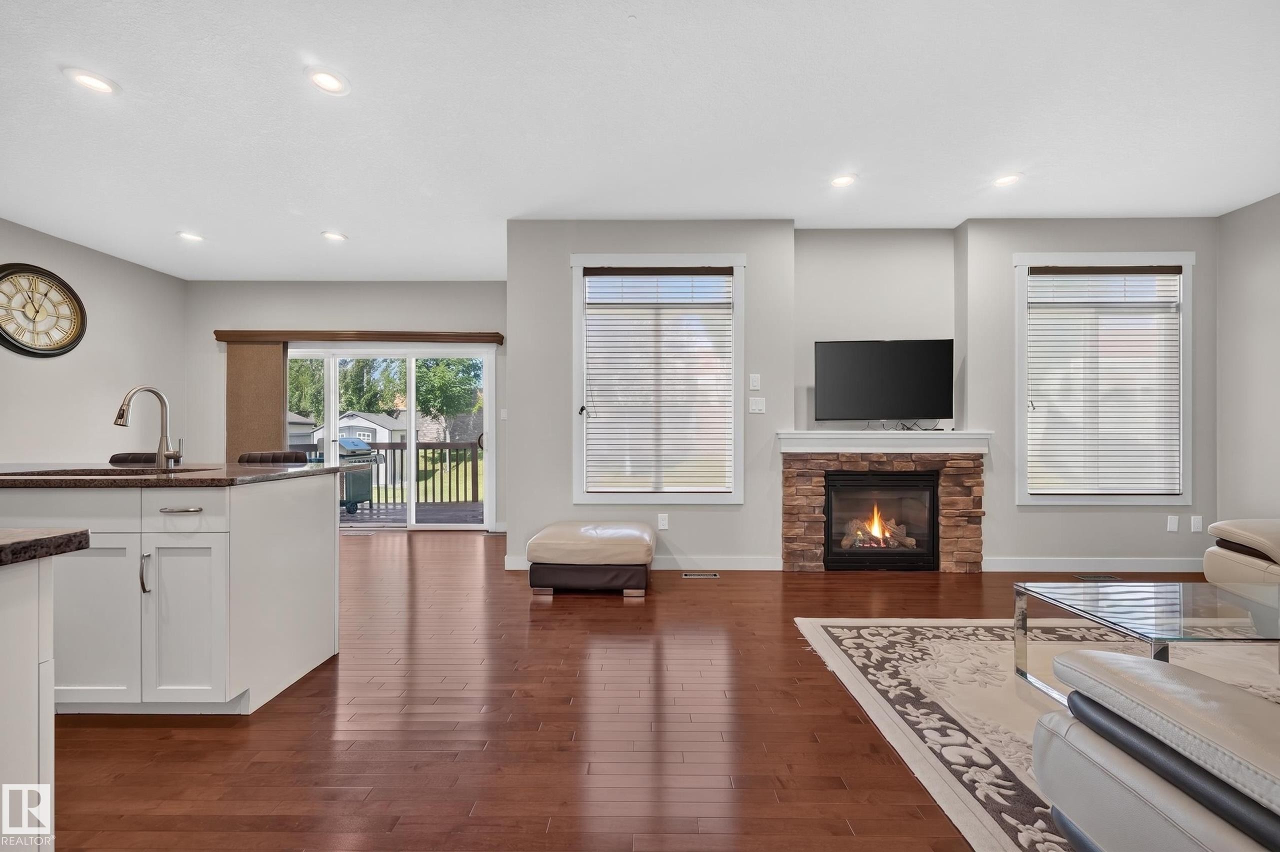 Open-concept living space featuring wood-finish flooring, a stone-faced fireplace with hearth, recessed lighting, and a sliding glass door leading to an exterior deck - 1512 Cunningham Cape, Edmonton, AB