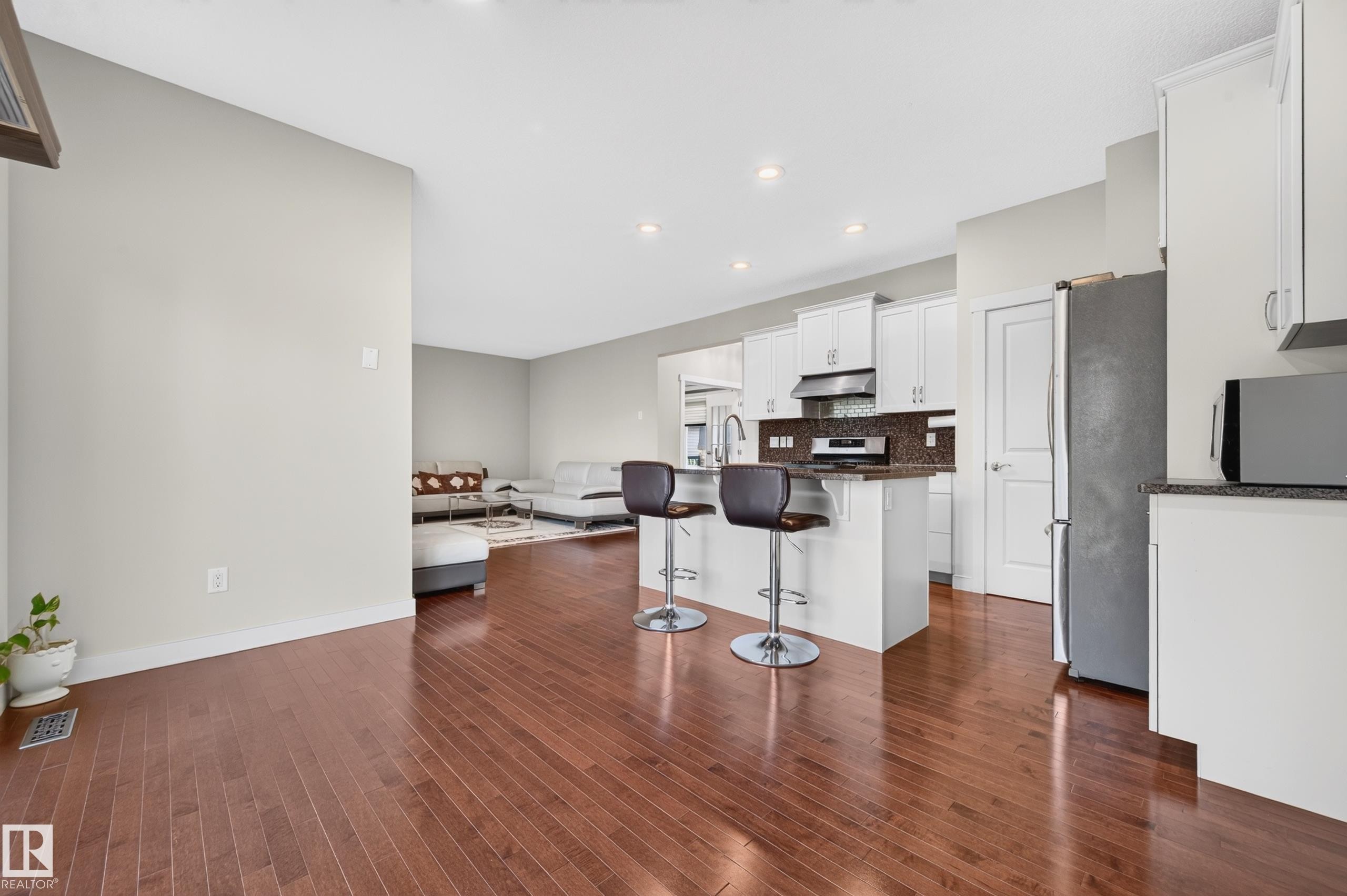Open-concept living space featuring wood-finish flooring, a kitchen island with seating, white cabinetry, dark countertops, and stainless steel appliances - 1512 Cunningham Cape, Edmonton, AB