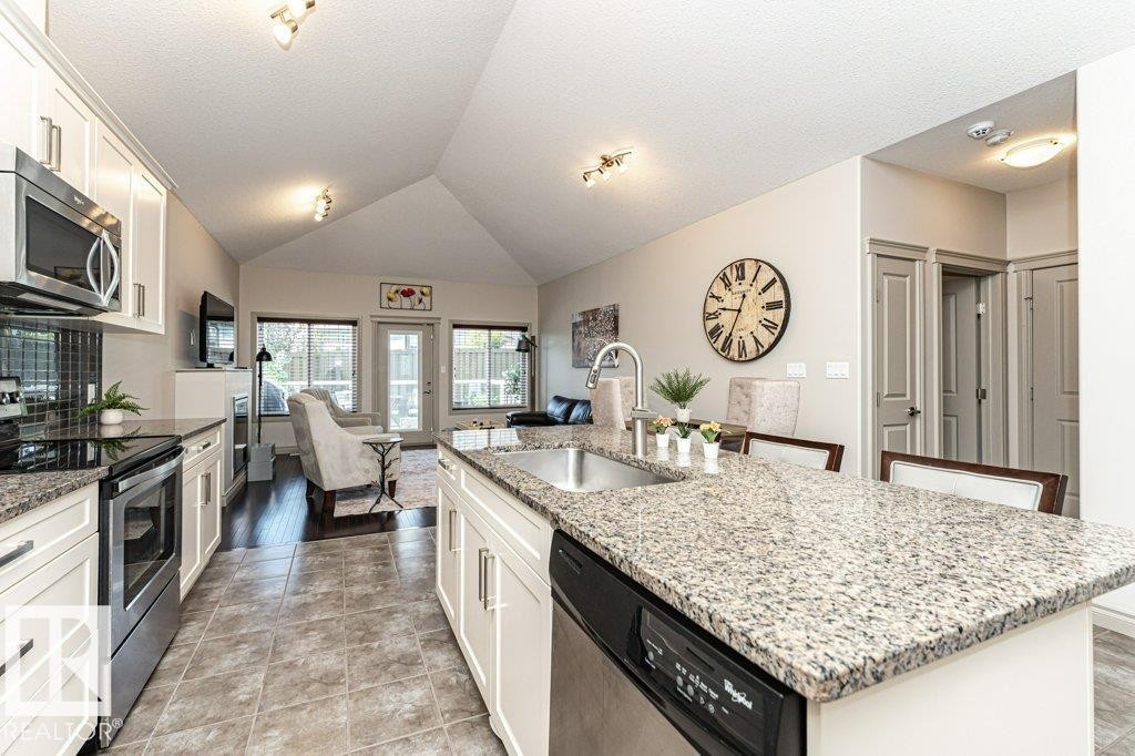 Open-concept kitchen featuring an island with an undermount sink, granite countertops, white cabinetry, and stainless steel appliances - 15 175 Mcconachie Drive, Edmonton, AB