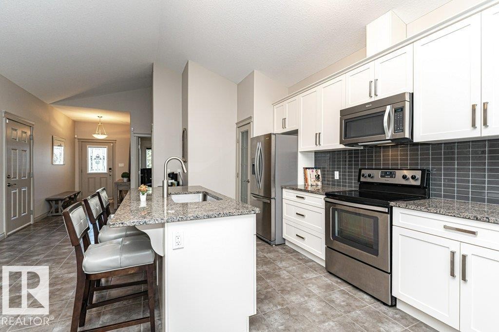 Kitchen featuring a central island with an undermount sink, granite countertops, white cabinetry, stainless steel appliances, and a gray subway tile backsplash - 15 175 Mcconachie Drive, Edmonton, AB