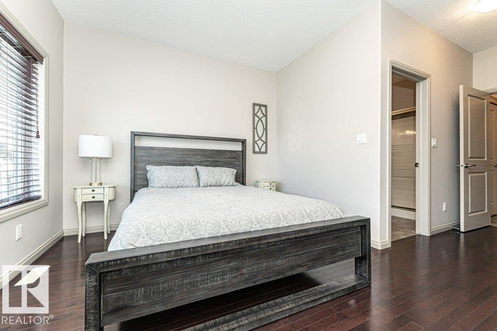 Bedroom featuring rich wood-finish flooring, light-toned walls, a window with horizontal blinds, and a doorway opening to a tiled shower enclosure - 15 175 Mcconachie Drive, Edmonton, AB