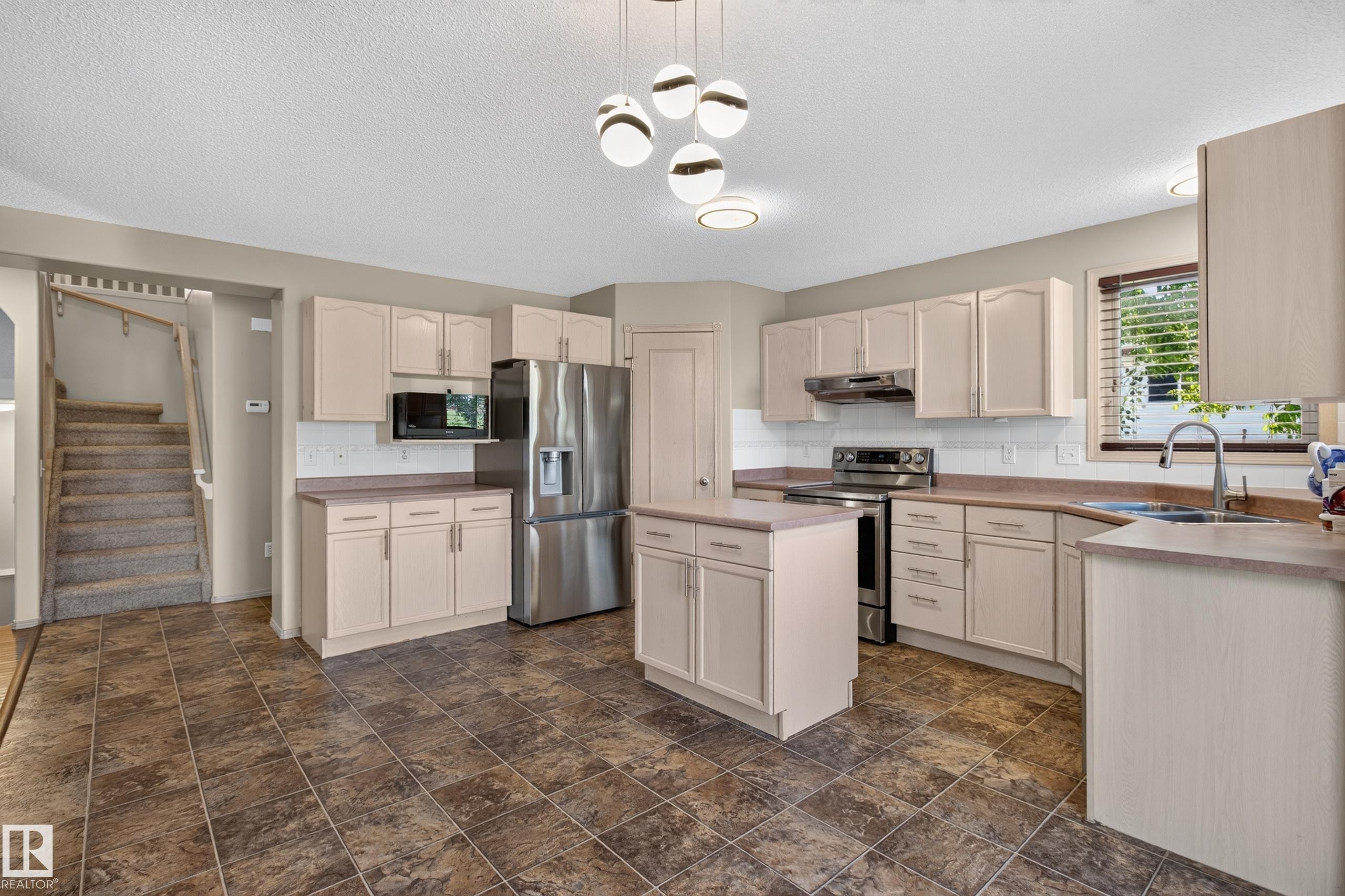 Kitchen featuring tile flooring, neutral cabinetry, a center island, stainless steel appliances, and a modern chandelier - 13406 32A Street, Edmonton, AB