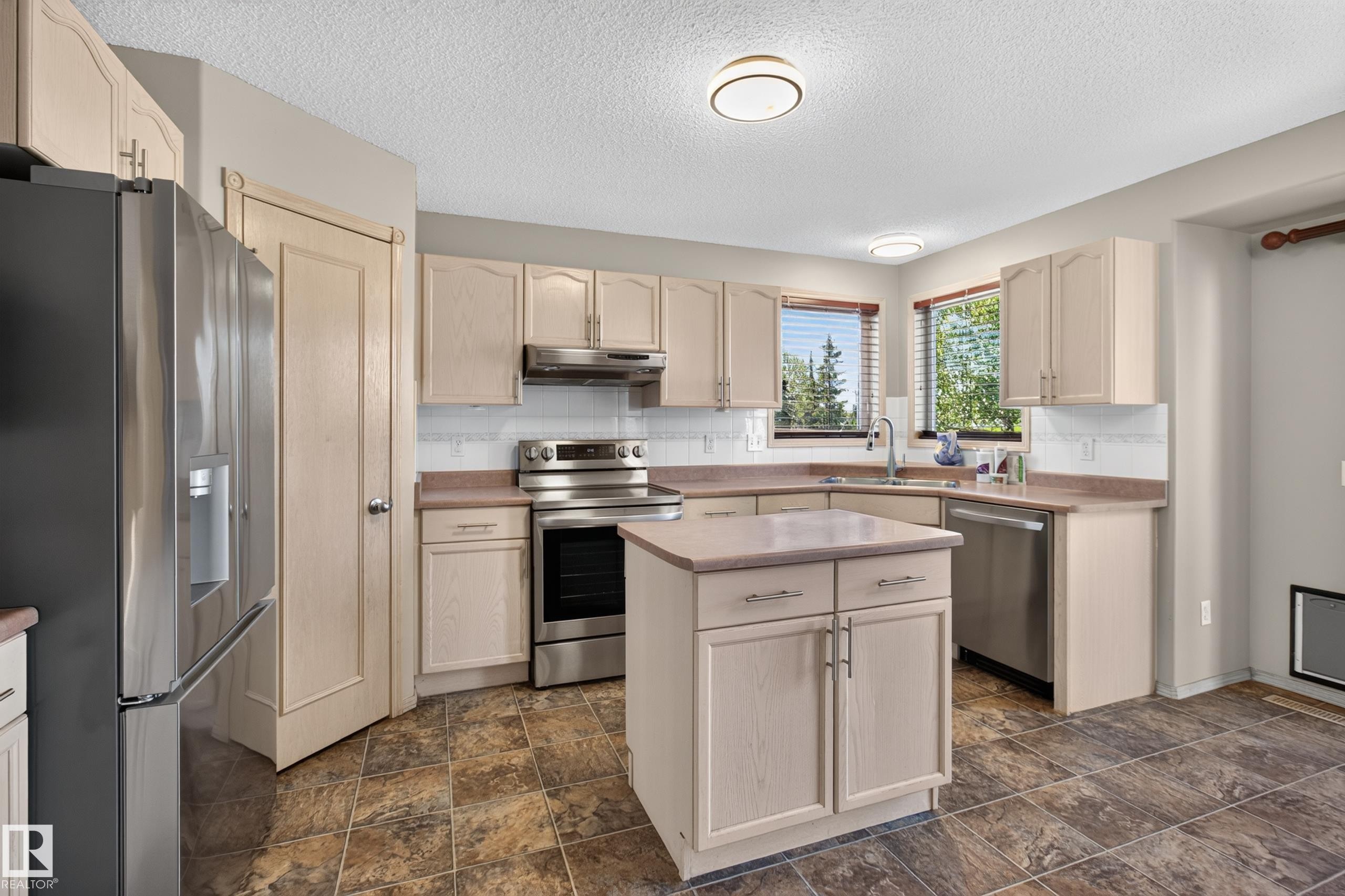 Kitchen featuring light-toned cabinetry, stainless steel appliances, a central island, tiled flooring, and dual windows - 13406 32A Street, Edmonton, AB