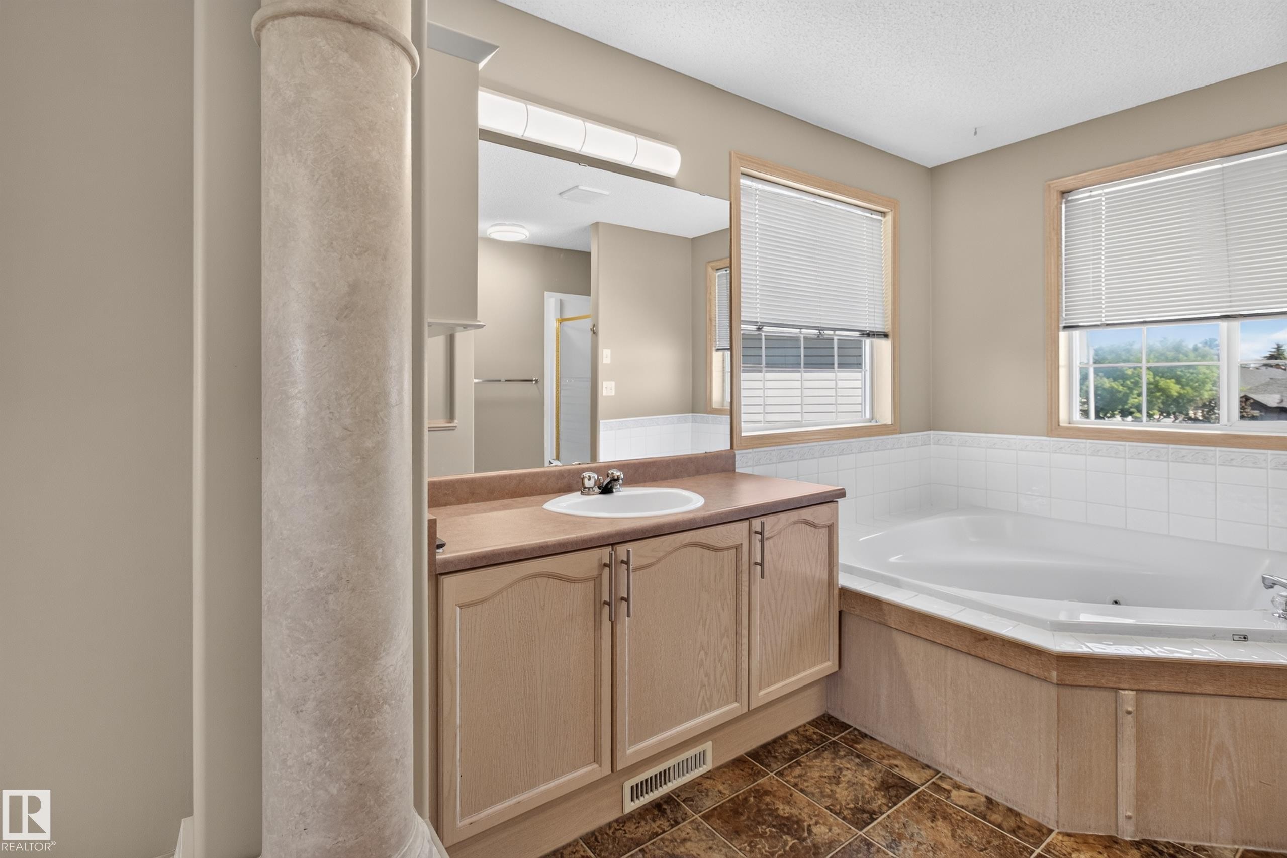 Bathroom featuring a built-in corner bathtub with white tile surround, a single vanity with a white basin sink and neutral countertop, two windows with blinds, and patterned floor tiles - 13406 32A Street, Edmonton, AB