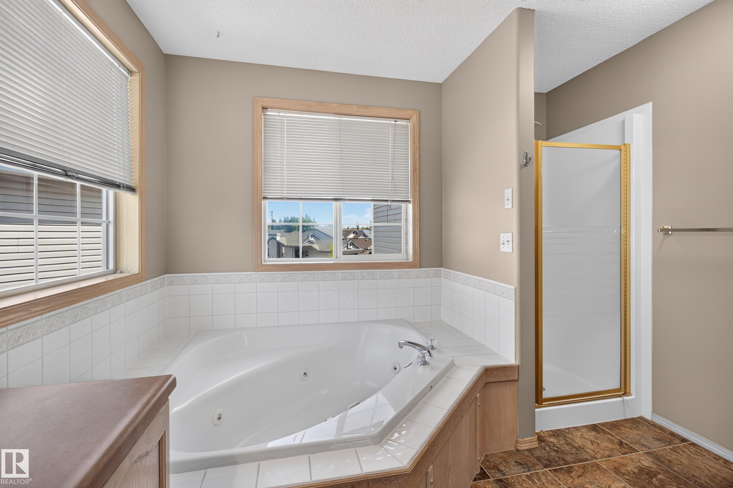 Corner jetted soaking tub with white tile surround, wood-finish trim, dual windows, and an adjacent shower enclosure with a gold-tone frame - 13406 32A Street, Edmonton, AB