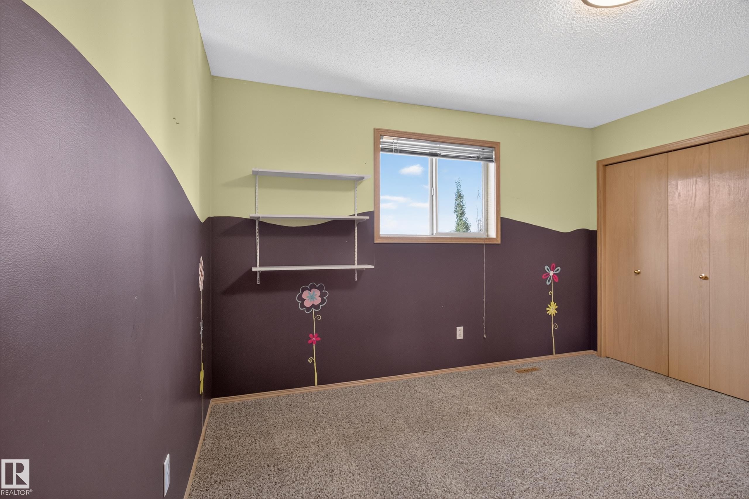 Carpeted room featuring a window with exterior visibility, light brown built-in bi-fold closet doors, and wall-mounted shelving - 13406 32A Street, Edmonton, AB