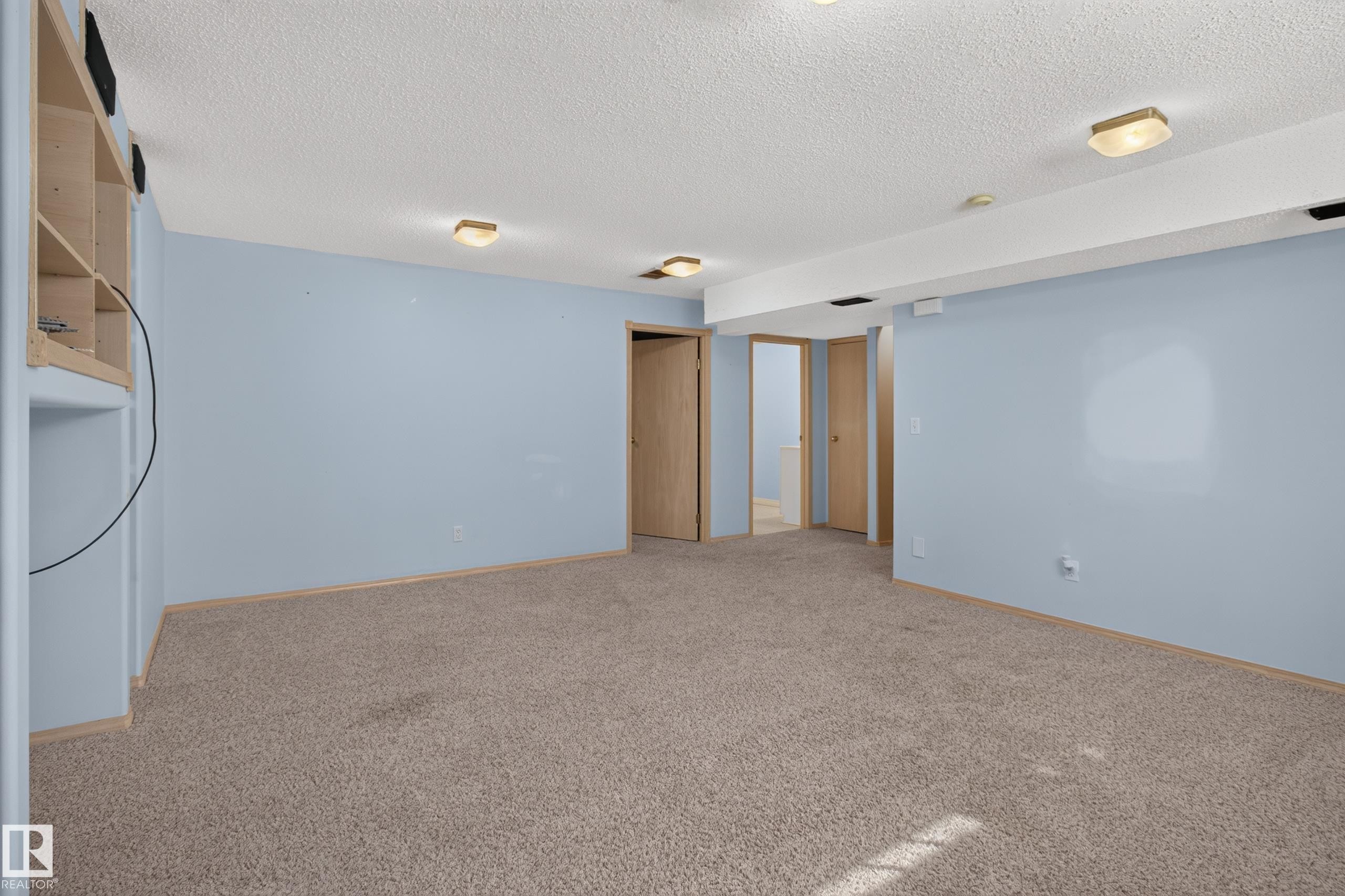 Spacious carpeted room featuring light blue walls, built-in shelving, multiple flush-mount light fixtures, and several interior doors - 13406 32A Street, Edmonton, AB