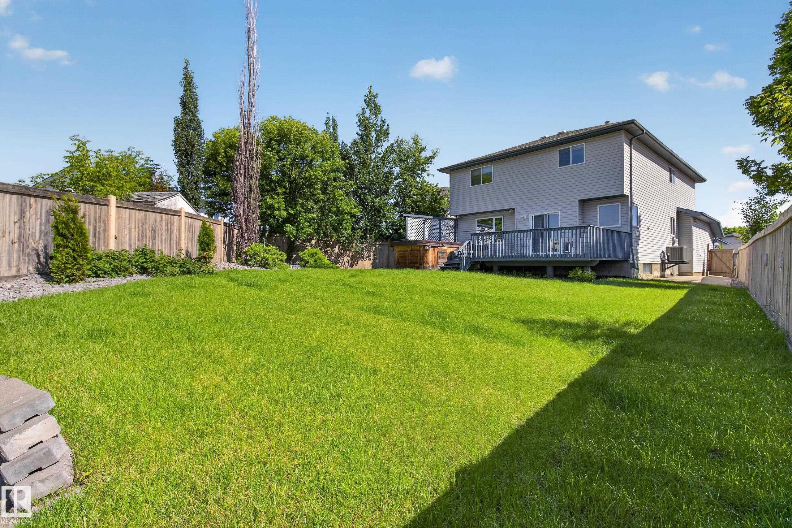 Expansive backyard featuring a lush green lawn, full privacy fencing, established perimeter landscaping, a two-story residence with light gray siding, and a spacious wooden deck - 13406 32A Street, Edmonton, AB