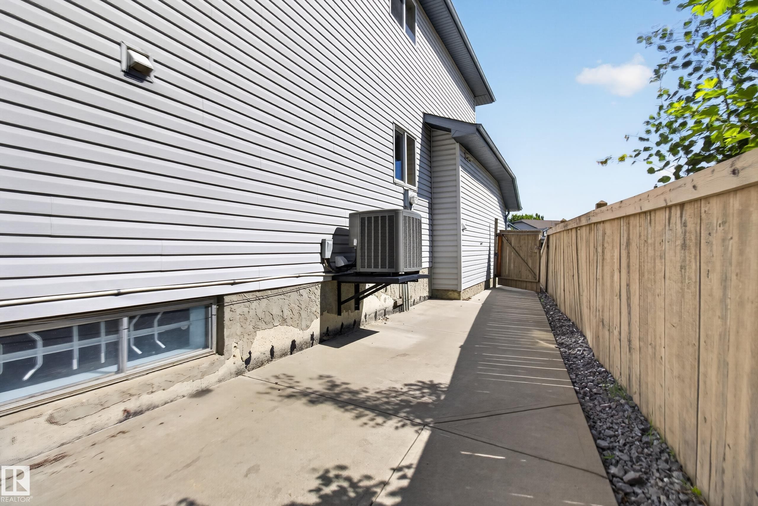 Concrete pathway alongside the property, featuring light-toned horizontal siding, a wooden privacy fence, a gate, and a visible exterior HVAC unit - 13406 32A Street, Edmonton, AB
