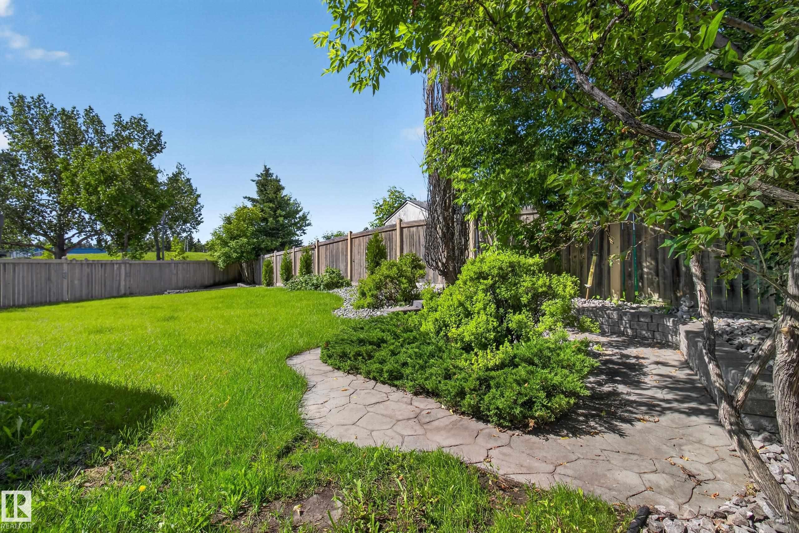 Expansive backyard featuring a lush lawn, stone pathway, established perimeter fencing, mature trees, and layered shrubbery - 13406 32A Street, Edmonton, AB