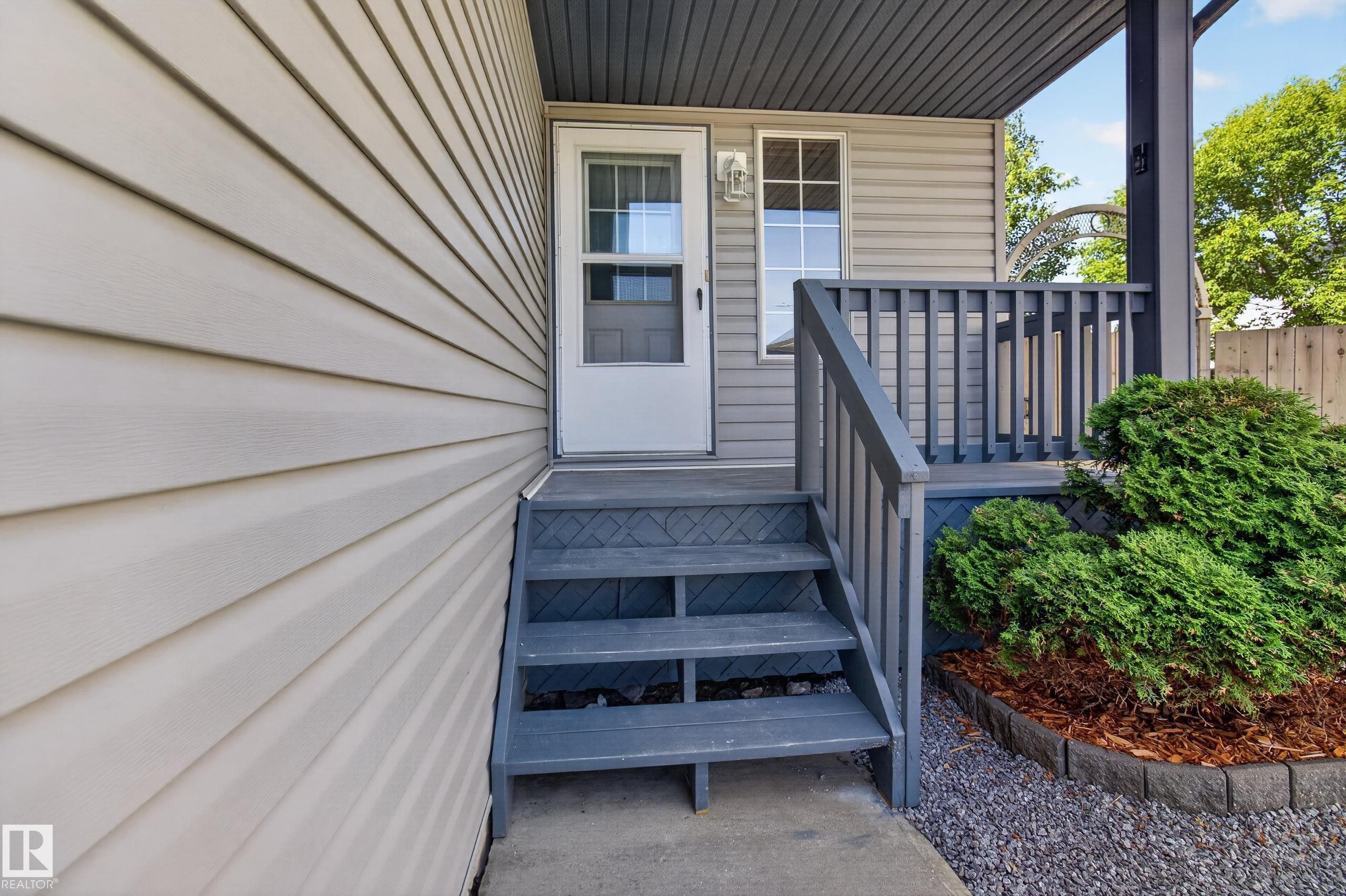 Front entry featuring a storm door with divided lites, exterior wall lantern, covered porch with dark gray railing, and steps leading to a concrete landing - 13406 32A Street, Edmonton, AB
