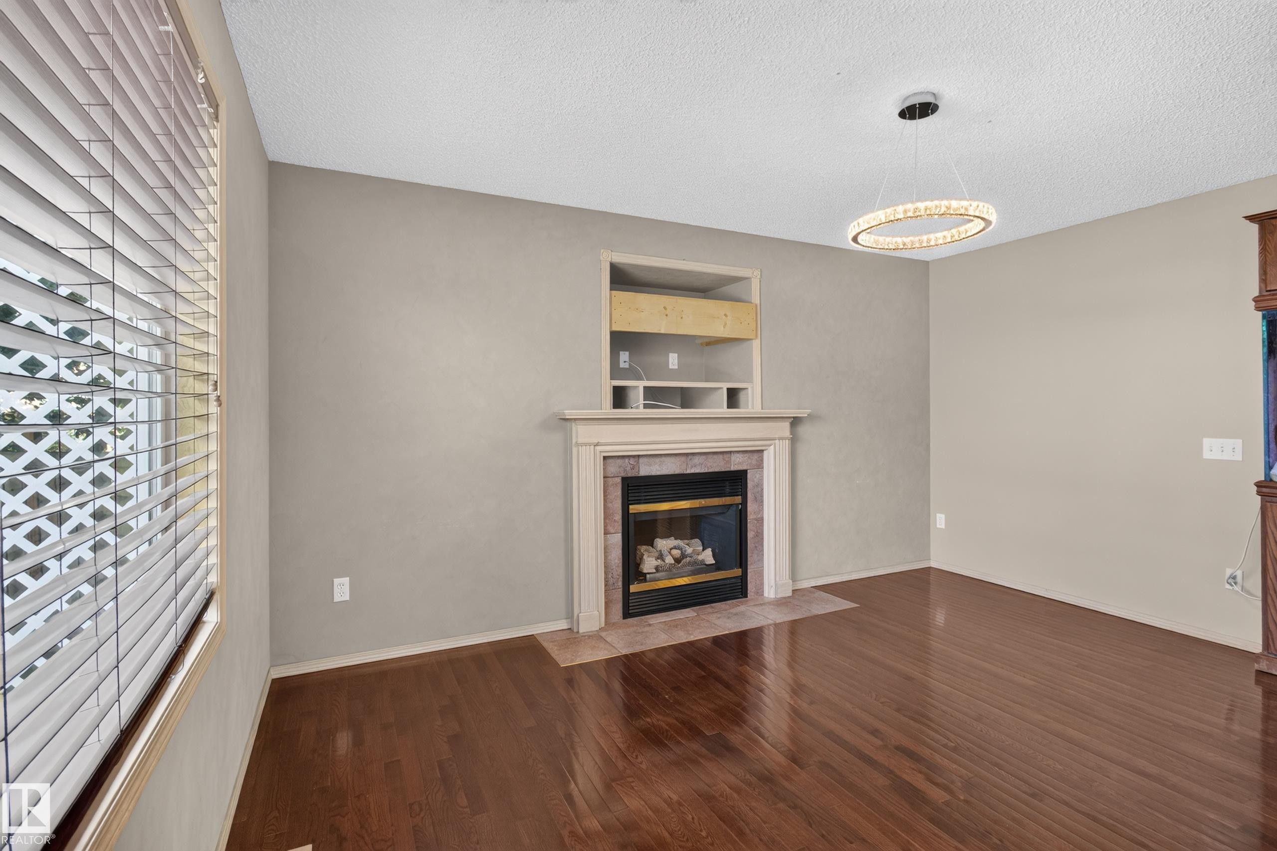 Living area featuring a fireplace with a stone surround and mantel, built-in shelving, wood-finish flooring, and a modern circular chandelier - 13406 32A Street, Edmonton, AB