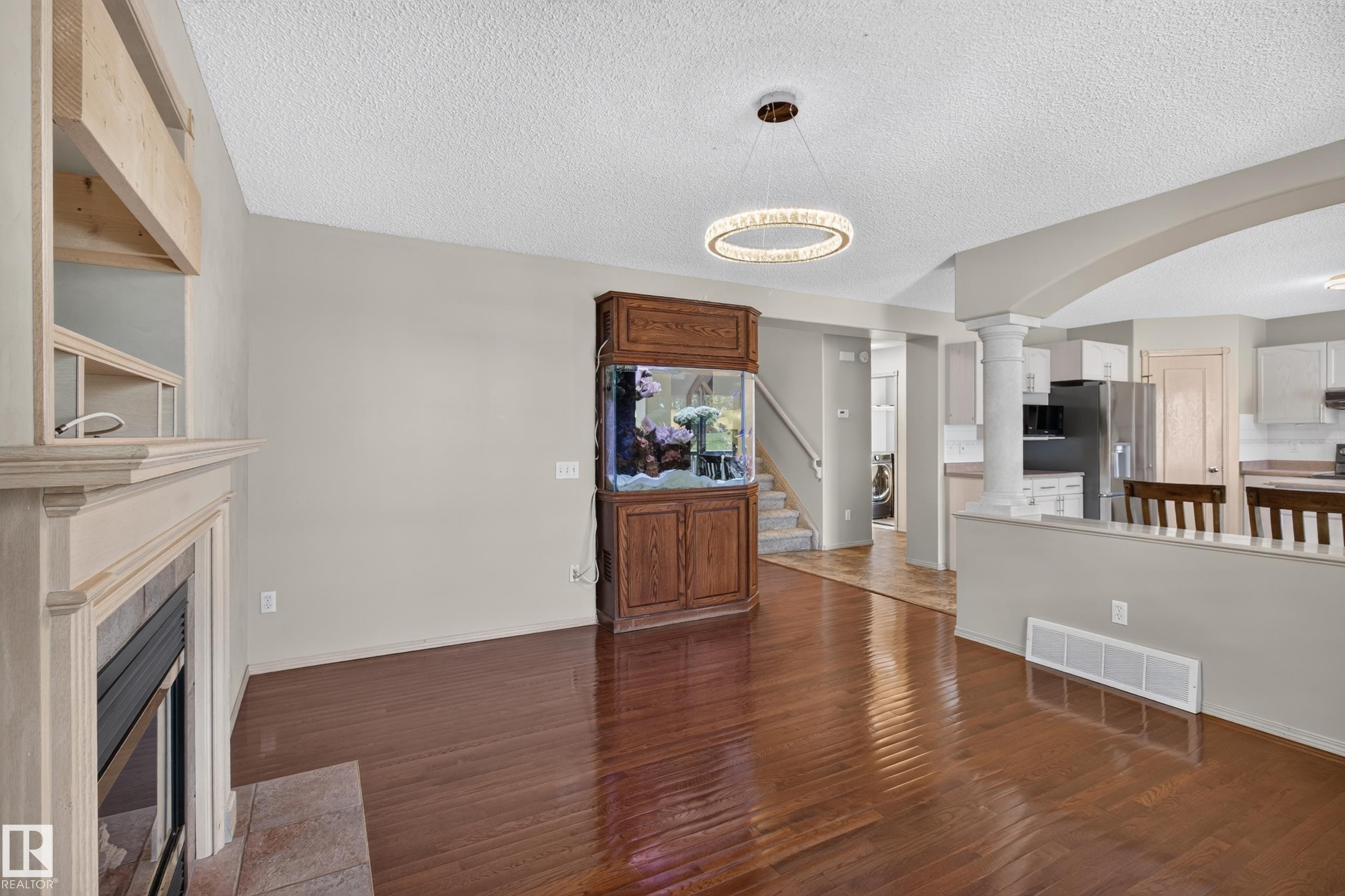 Expansive main living area featuring wood-finish flooring, a contemporary chandelier, a built-in fireplace with a tiled hearth, and a partial wall with an arched opening to the kitchen - 13406 32A Street, Edmonton, AB