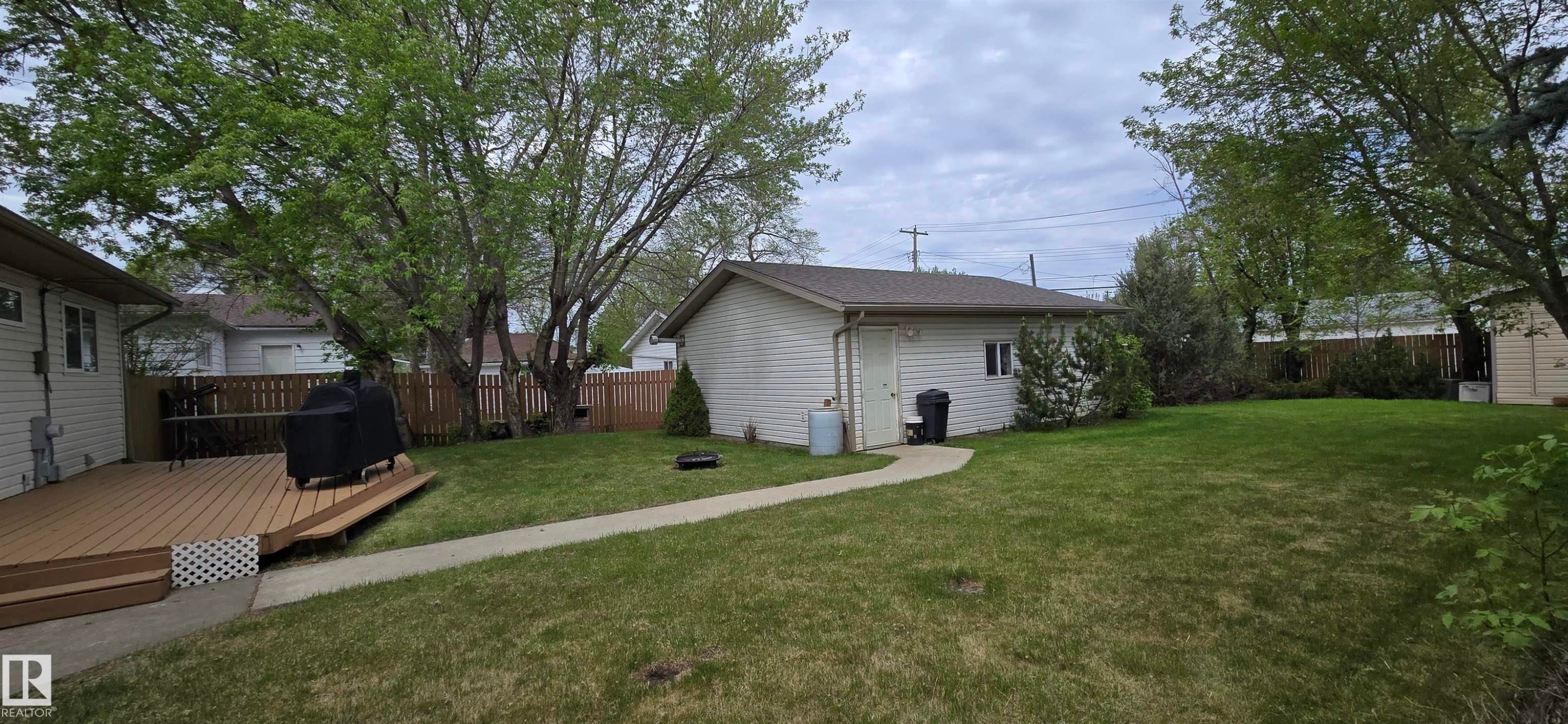 Expansive backyard featuring a manicured lawn, an elevated wood-finish deck, a concrete pathway, a detached outbuilding, and mature trees - 4734 54A Avenue, Vegreville, AB - Outdoor
