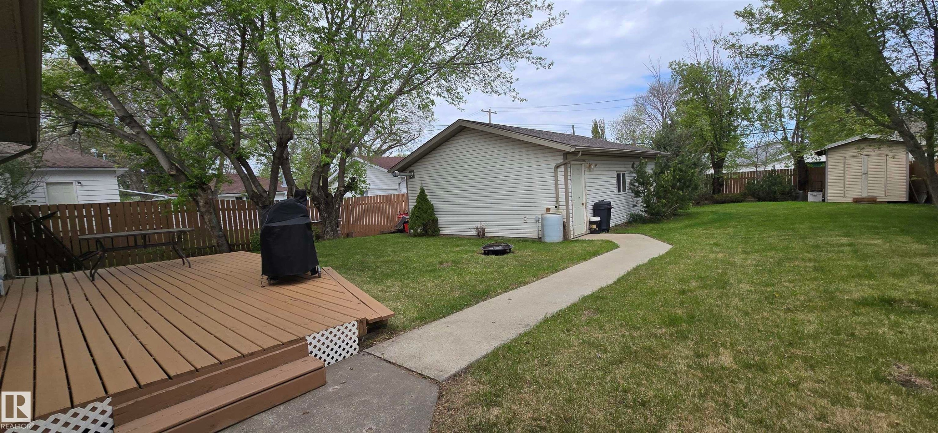Expansive backyard featuring a large wood-finish deck with lattice skirting, a concrete pathway, a detached single-story structure with siding, a separate storage shed, and mature trees - 4734 54A Avenue, Vegreville, AB - Outdoor