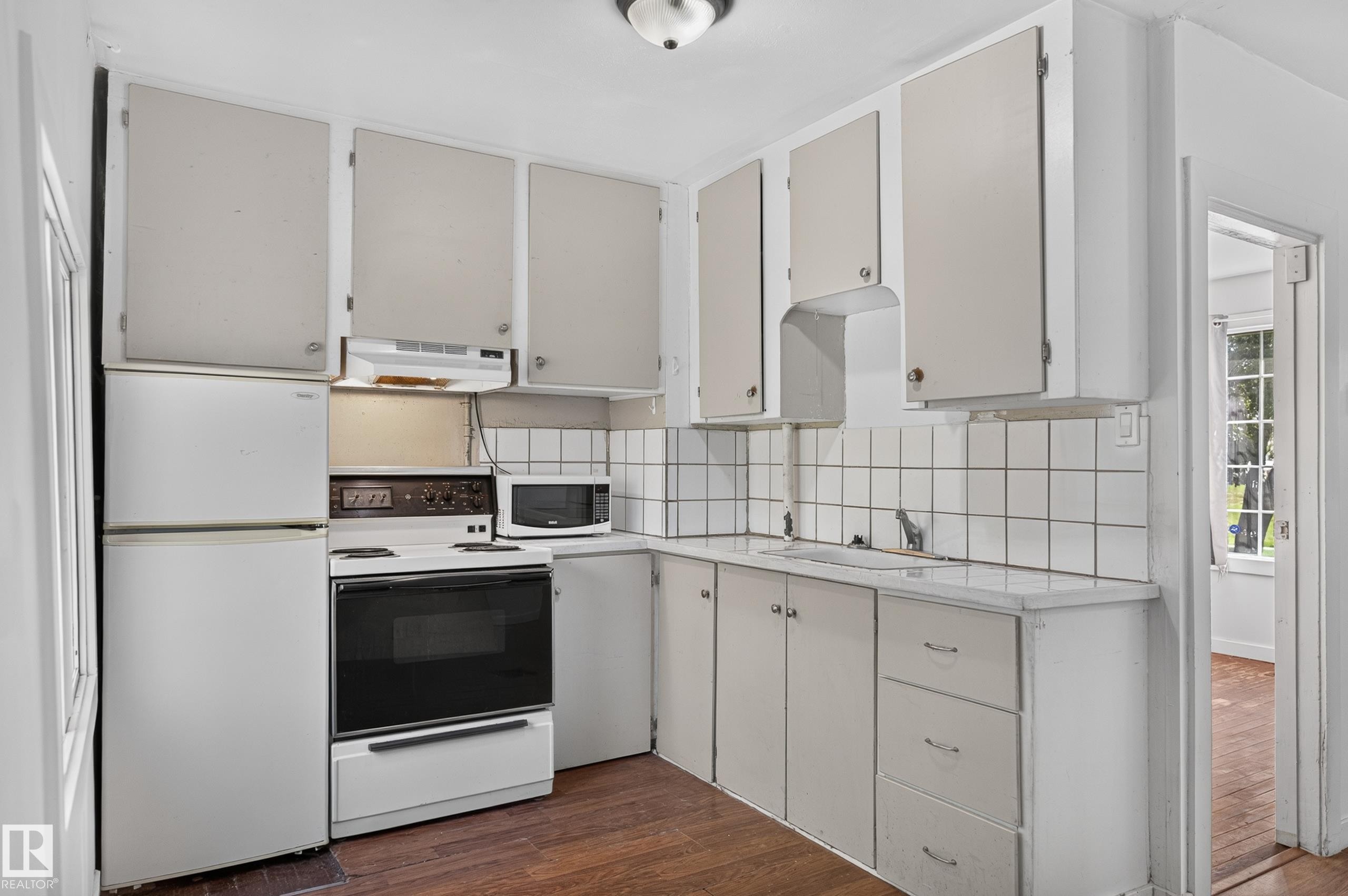 Compact kitchen featuring white cabinetry, a built-in oven with cooktop, a white refrigerator, and a microwave - 11708 79 Street, Edmonton, AB