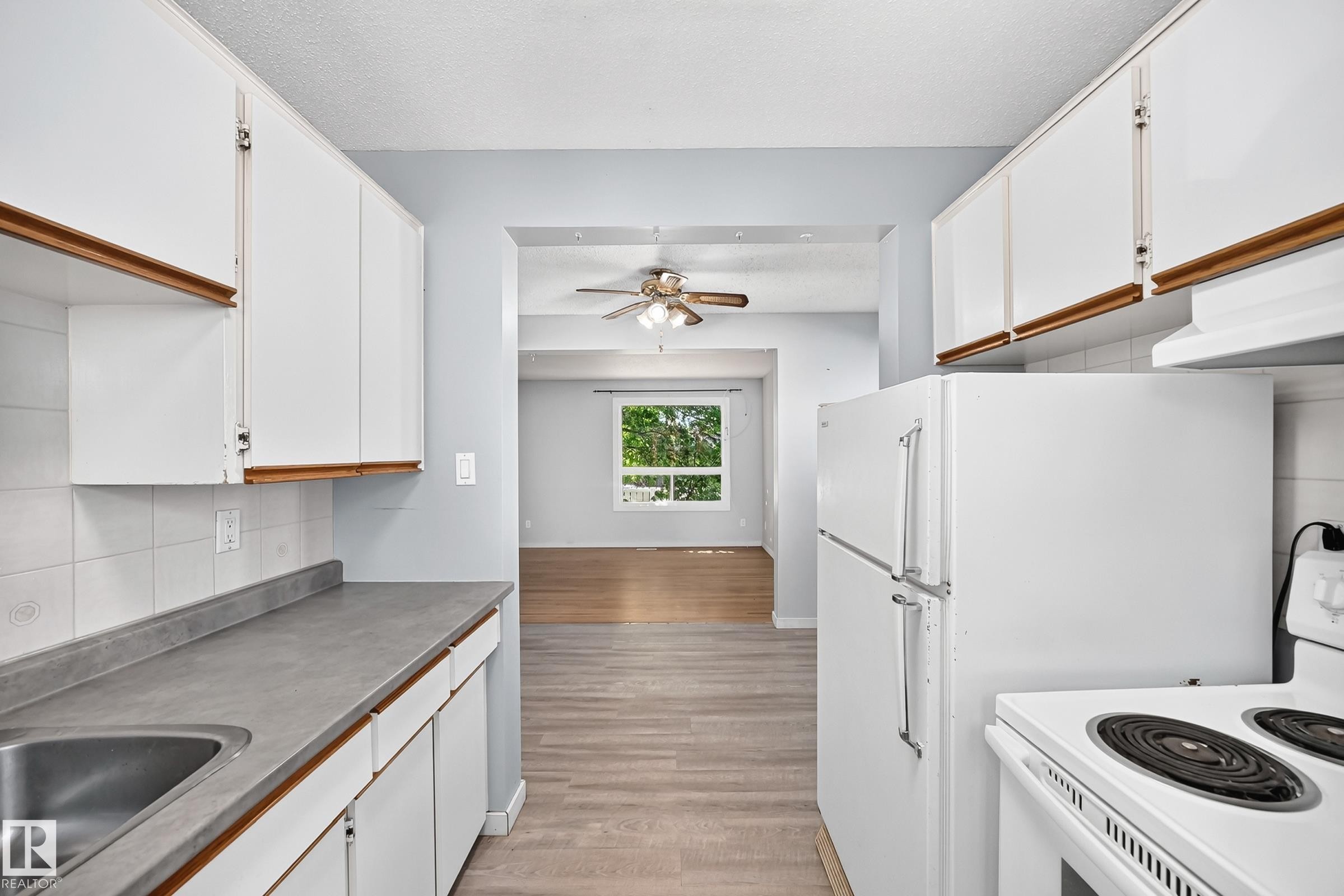 Functional kitchen featuring white cabinetry, a stainless steel sink, and white appliances, with an adjacent room offering wood-finish flooring and a ceiling fan - 355 Northgate Terrace, Edmonton, AB