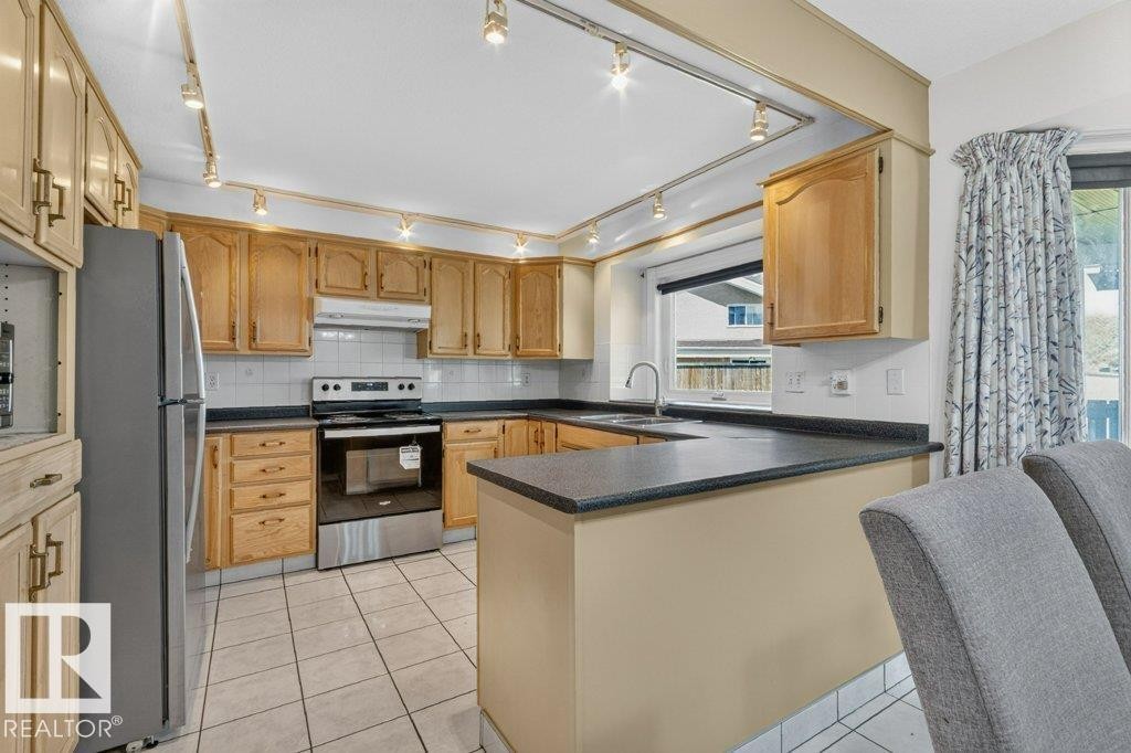 U-shaped kitchen featuring wood cabinetry, dark countertops, white tile backsplash, and tile flooring - 16524 99 Street Nw, Edmonton, AB - Indoor Photo Showing Kitchen