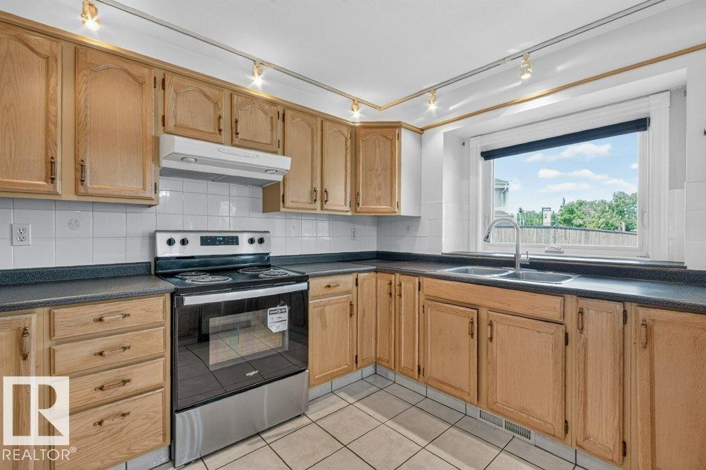 Kitchen featuring light wood-finish cabinetry, dark countertops, white tile backsplash, a stainless steel range, and a double-basin stainless steel sink under a window - 16524 99 Street Nw, Edmonton, AB - Indoor Photo Showing Kitchen With Double Sink