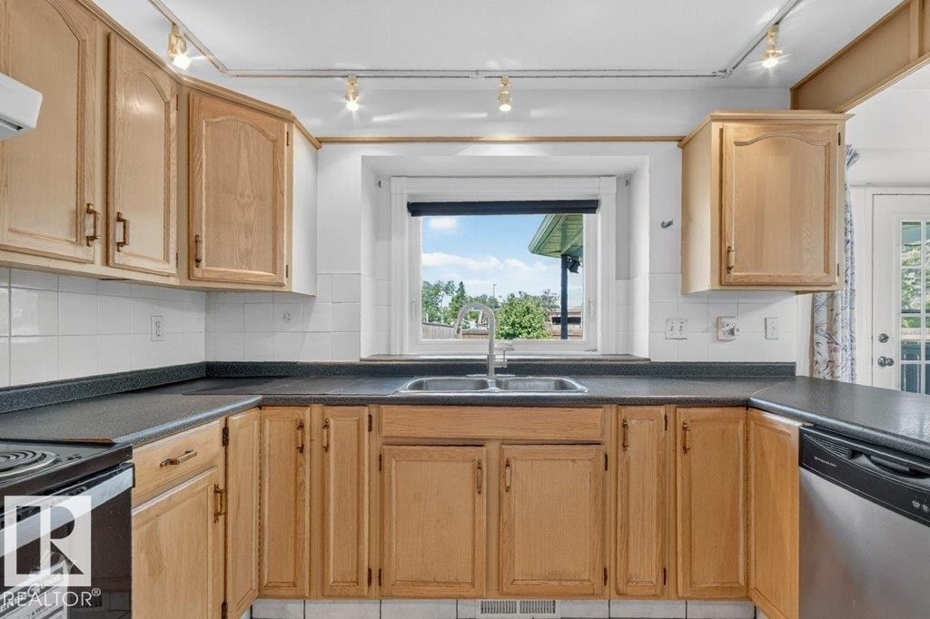 Functional kitchen featuring light wood cabinetry, dark countertops, a double basin stainless steel sink, and white subway tile backsplash - 16524 99 Street Nw, Edmonton, AB - Indoor Photo Showing Kitchen With Double Sink