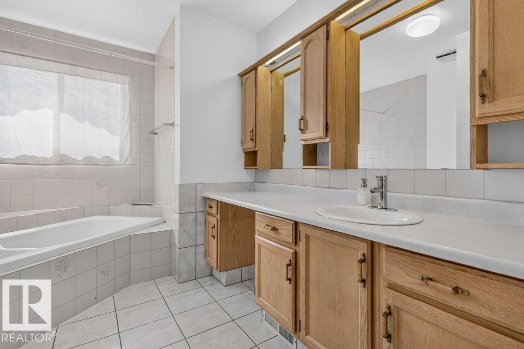 Bathroom featuring a built-in tub with white tile surround, a spacious vanity with light wood cabinetry, a white countertop, and an integrated sink with chrome faucet - 16524 99 Street Nw, Edmonton, AB - Indoor Photo Showing Bathroom