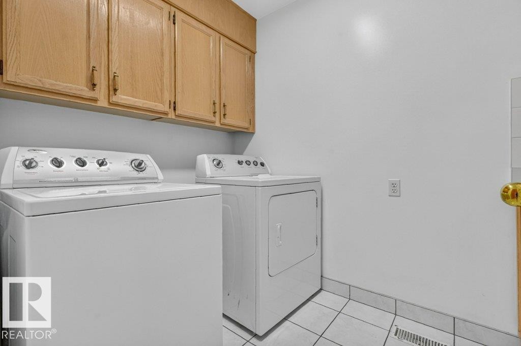 Dedicated main floor laundry area featuring a white top-load washer, a white dryer, wood-finish upper cabinetry, white walls, and a tiled floor - 16524 99 Street Nw, Edmonton, AB - Indoor Photo Showing Laundry Room