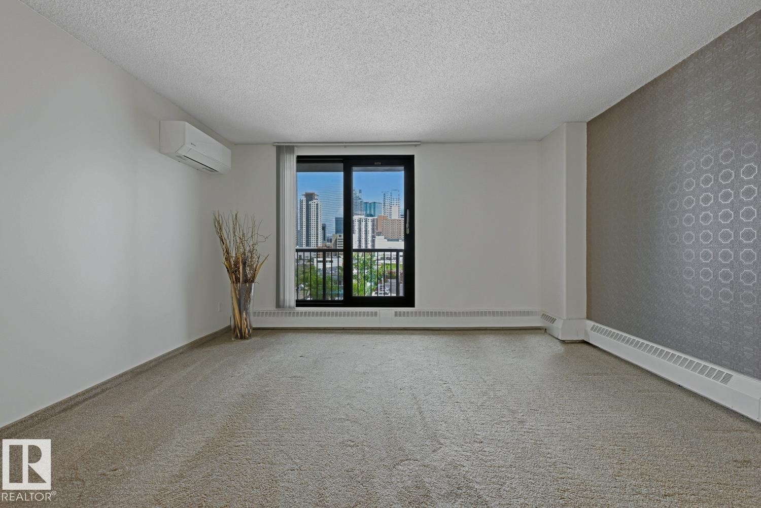 Carpeted interior space featuring a full-height glass door with an integrated balcony, a cityscape backdrop, a ductless mini-split air conditioning unit, patterned accent wall, and baseboard heating - 505 10145 109 Street, Edmonton, AB