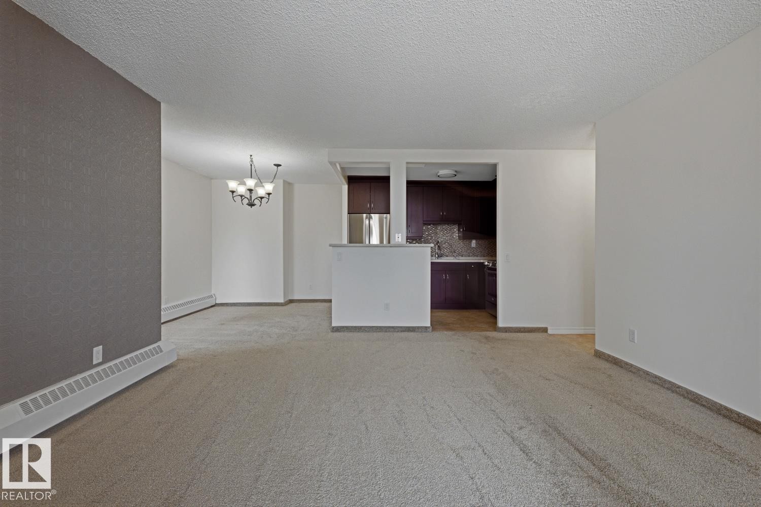 Open concept living area with light-colored carpeting, a partial wall separating the kitchen, and a dining area featuring a chandelier - 505 10145 109 Street, Edmonton, AB