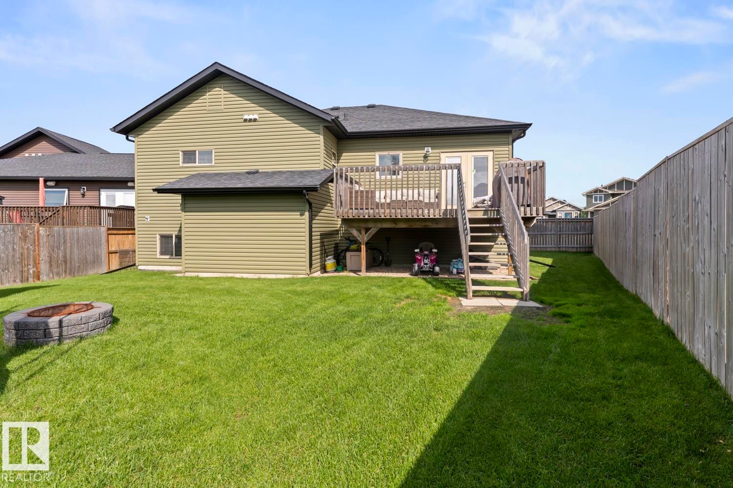 Expansive backyard featuring a manicured lawn, a built-in fire pit, a wooden deck with stairs, a privacy fence, and light green vinyl siding - 35 Falcon Road, Cold Lake, AB