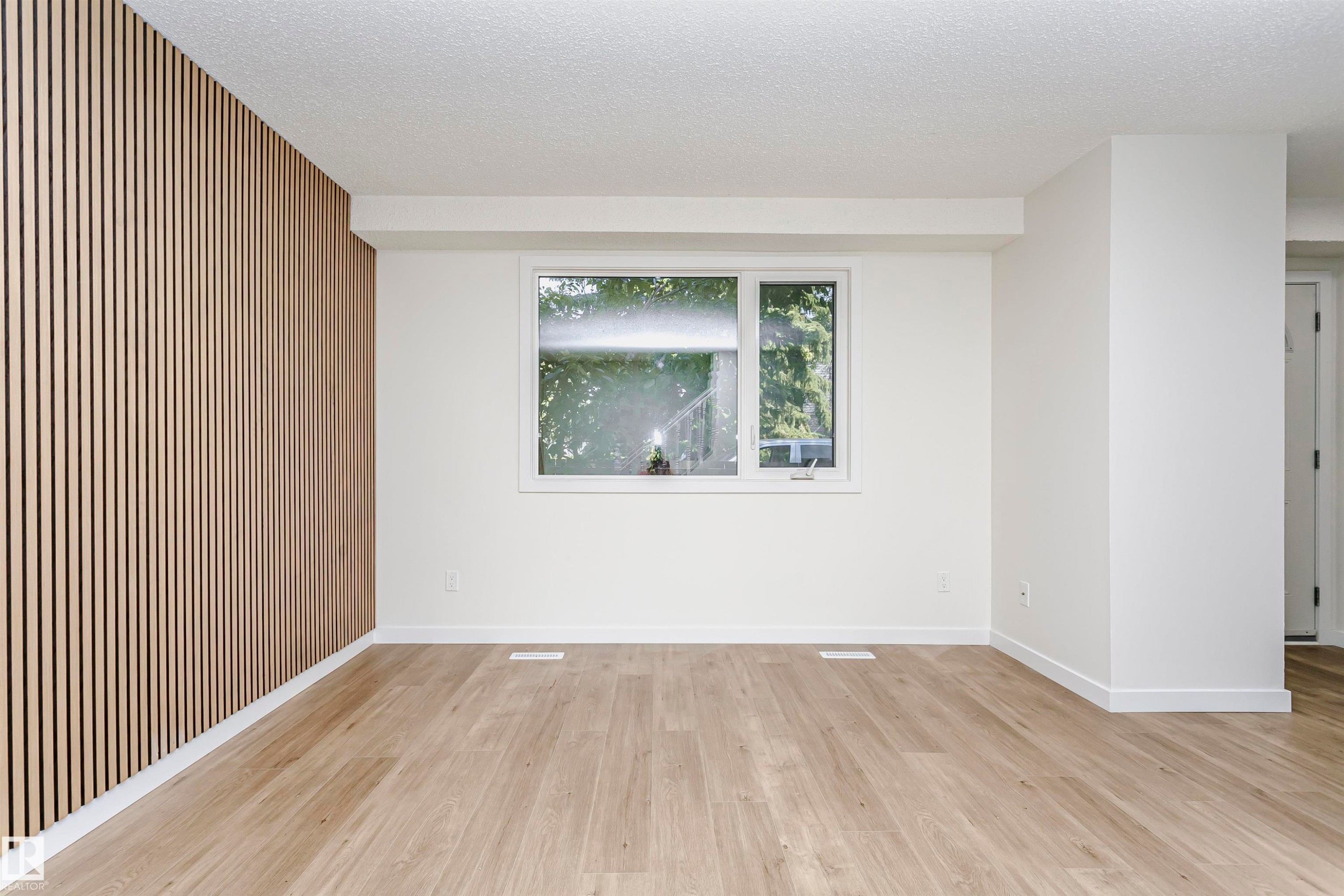 Spacious interior featuring a contemporary accent wall with vertical wood paneling, light wood-finish flooring, and a large window providing natural light - 5418 144B Avenue, Edmonton, AB