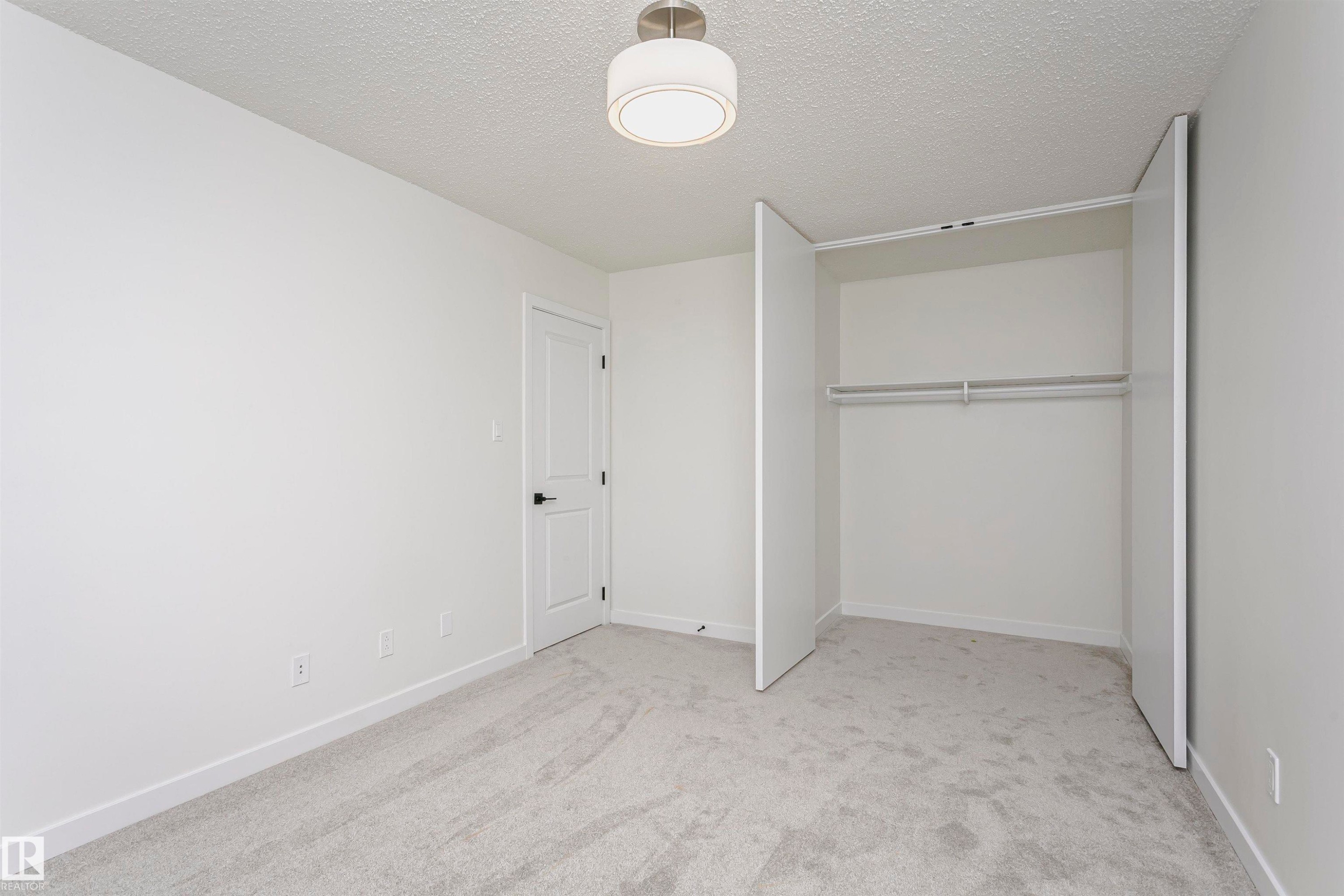 Bedroom featuring light gray carpet flooring, white walls, a flush-mount ceiling light fixture, a closet with bi-fold doors, and a solid interior door with black hardware - 5418 144B Avenue, Edmonton, AB