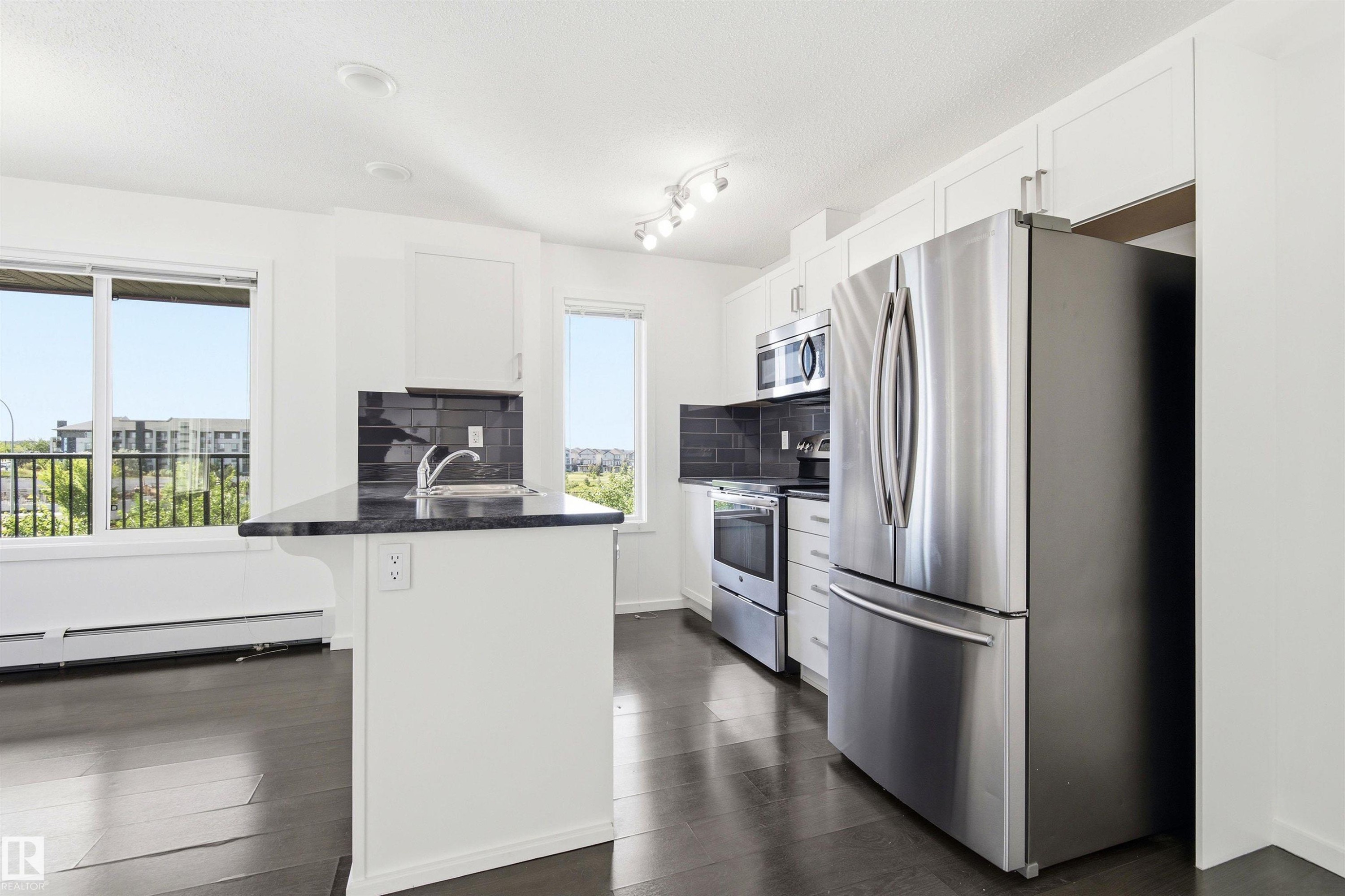 Kitchen featuring white cabinetry, stainless steel appliances, dark countertops, a dark tile backsplash, and dark wood-finish flooring - 435 5515 7 Avenue, Edmonton, AB