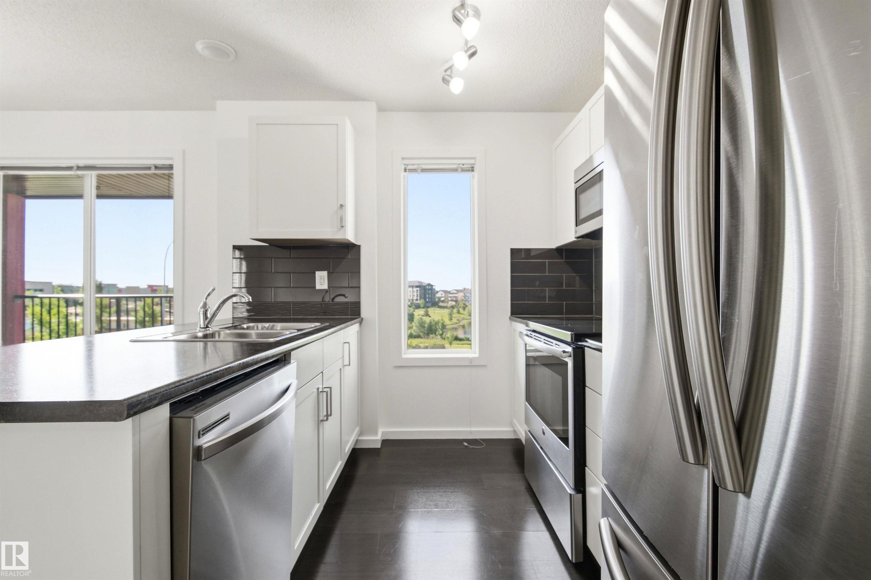Modern kitchen featuring stainless steel appliances, dark wood-finish flooring, white cabinetry, dark subway tile backsplash, and dark countertops - 435 5515 7 Avenue, Edmonton, AB
