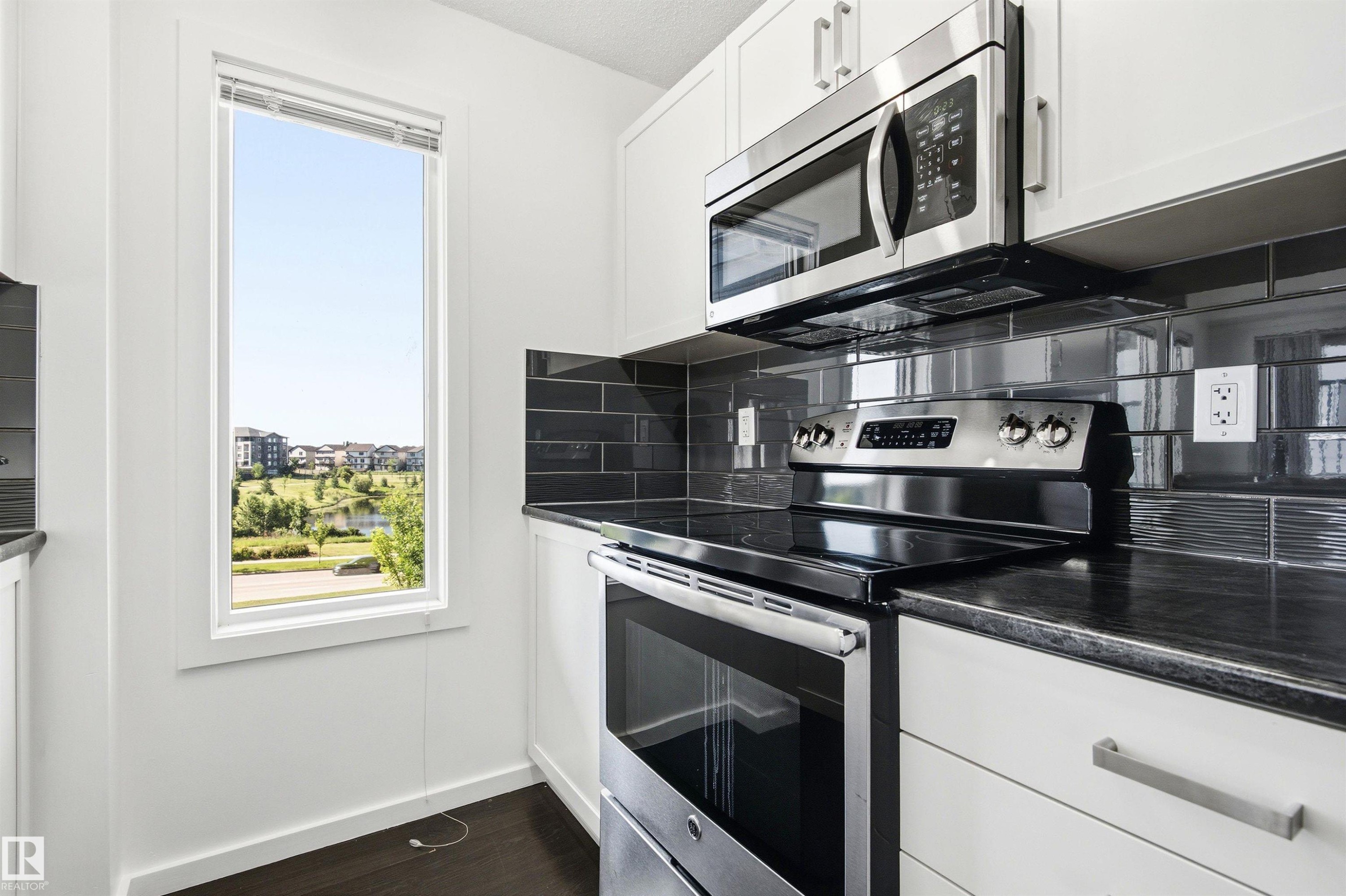 Modern kitchen featuring white cabinetry, dark countertop, stainless steel appliances, dark subway tile backsplash, and a tall window - 435 5515 7 Avenue, Edmonton, AB