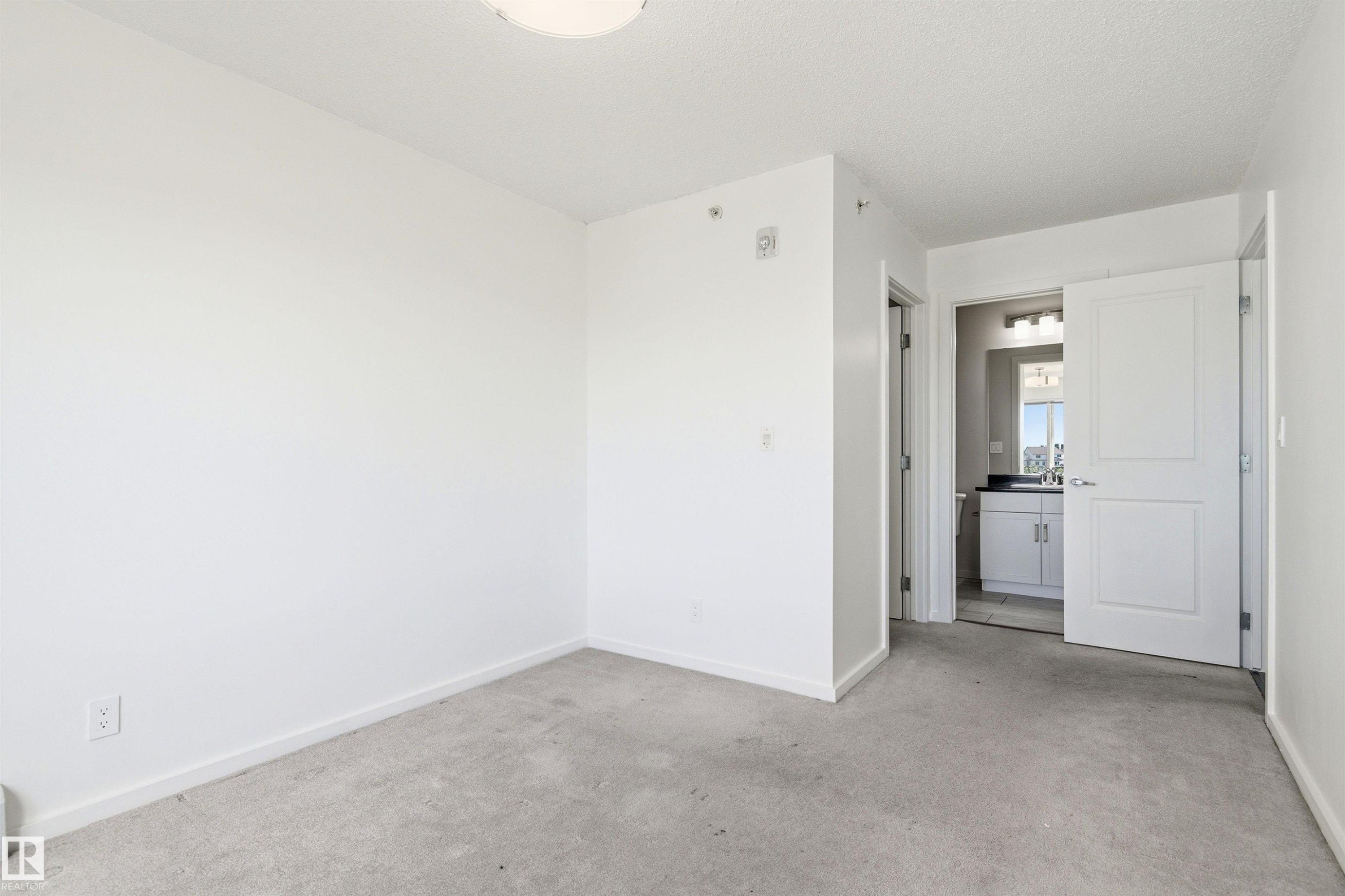 Carpeted room featuring a white ceiling and baseboards, with a bathroom entrance revealing a vanity with a dark countertop and mirror - 435 5515 7 Avenue, Edmonton, AB