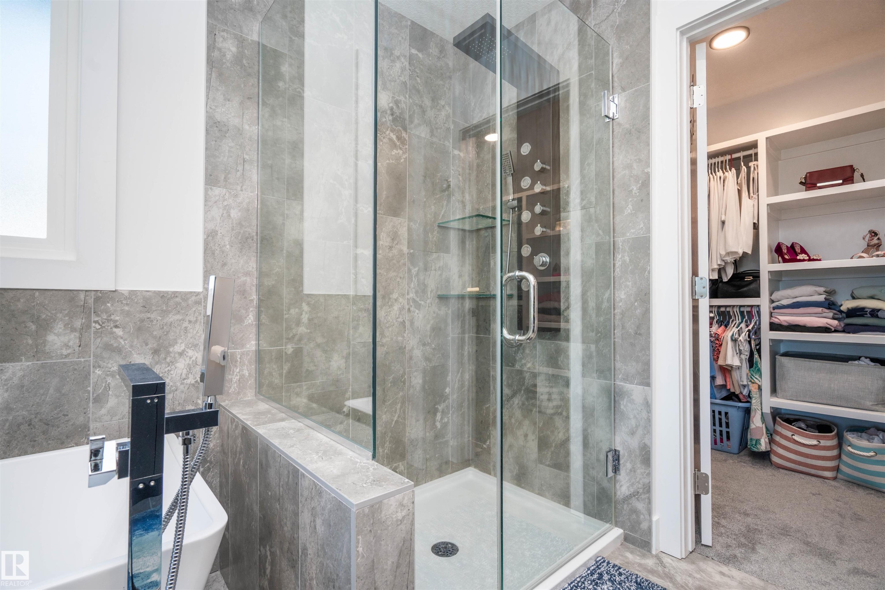 Contemporary bathroom featuring a frameless glass shower enclosure with rain shower head, full-height stone-finish wall tiling, separate soaking tub with a modern floor-mounted fixture, and a built-in walk-in closet with integrated shelving - 4714 Charles Bay Bay, Edmonton, AB