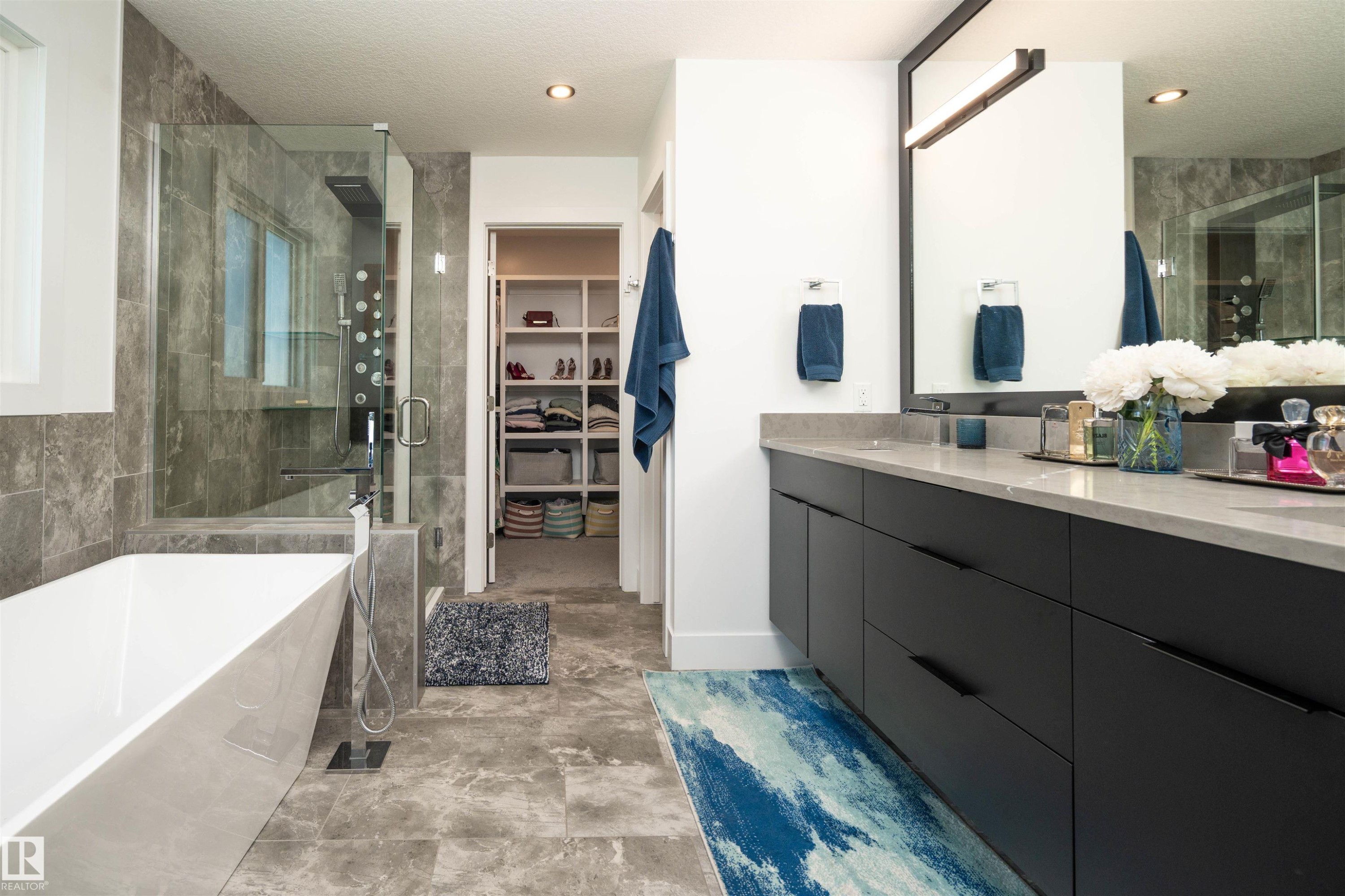 Contemporary bathroom featuring a freestanding soaking tub with a floor-mounted faucet, a glass-enclosed shower with multiple sprayers, a double vanity with dark cabinetry, and stone-look tile flooring - 4714 Charles Bay Bay, Edmonton, AB