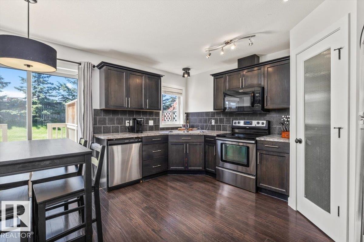Kitchen featuring dark wood-finish cabinetry, stainless steel appliances, dark tile backsplash, and wood-finish flooring - 125 Birchwood Close, Devon, AB