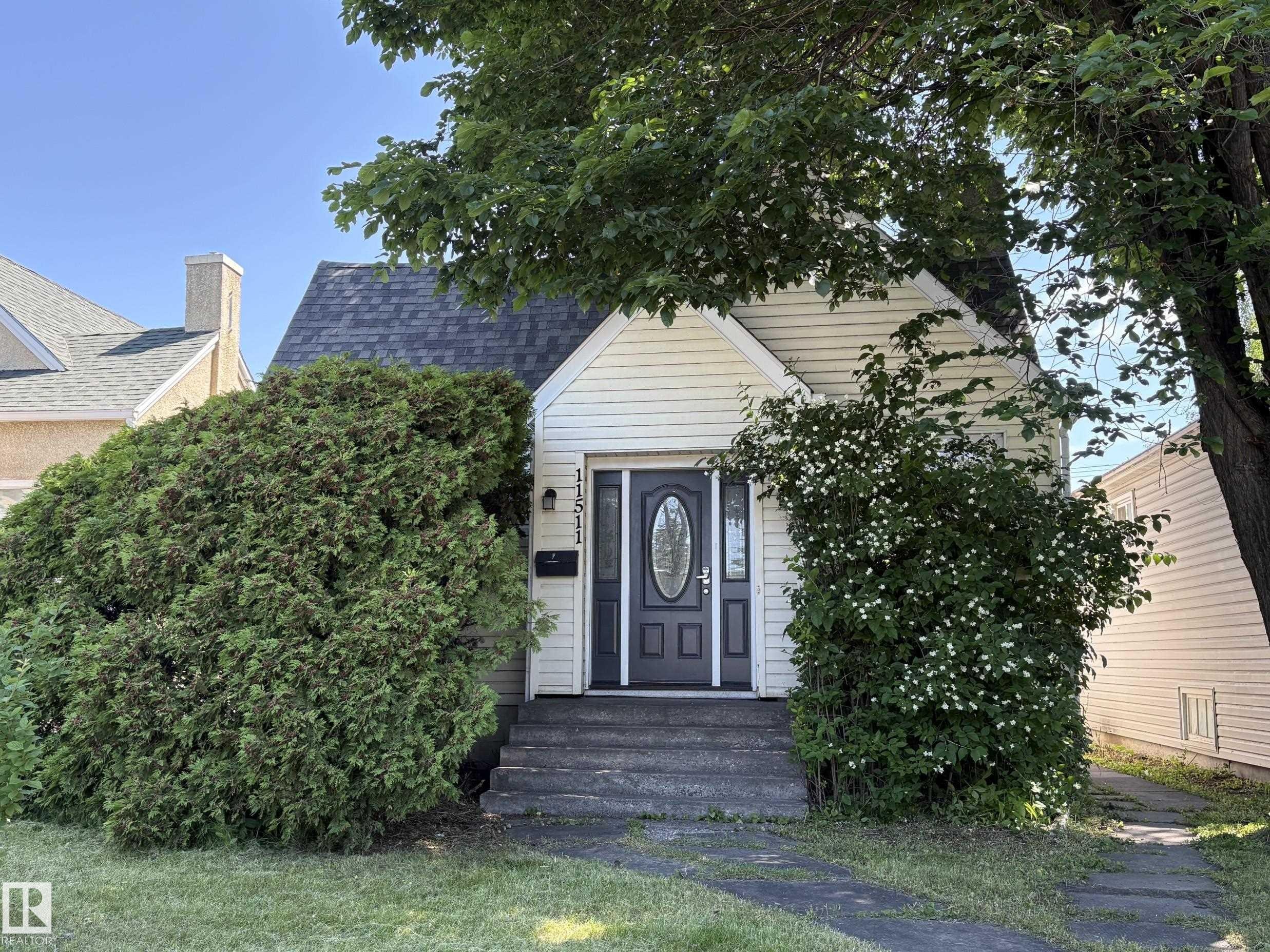Charming cottage-style home featuring light-colored siding, a dark shingles roof, and an inviting front entrance with a dark door and clear glass insert - 11511 101 Street, Edmonton, AB
