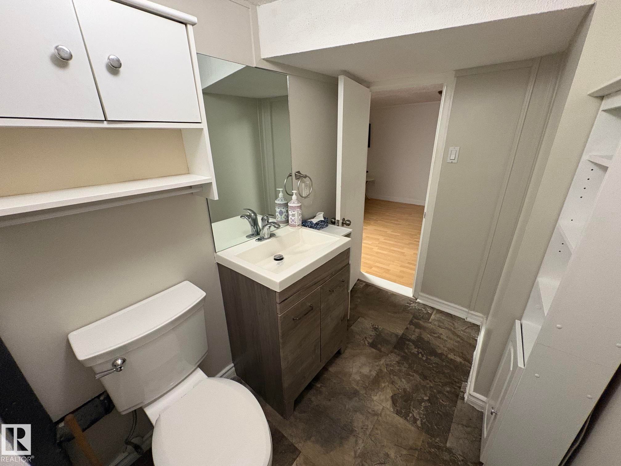 Bathroom featuring a vanity with a white basin and chrome faucet, a wall-mounted mirror, overhead cabinetry, and a toilet - 11511 101 Street, Edmonton, AB