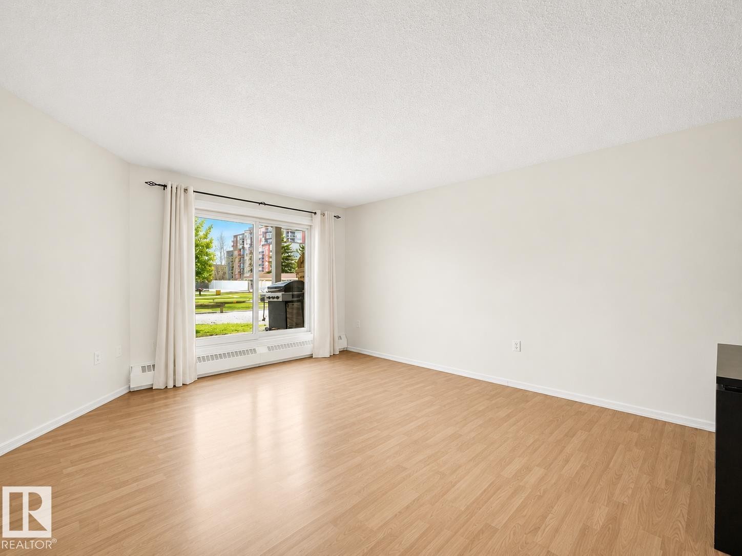 Spacious interior featuring wood-finish flooring and a large window with white curtain panels - #143 10636 120 St Nw, Edmonton, AB - Indoor