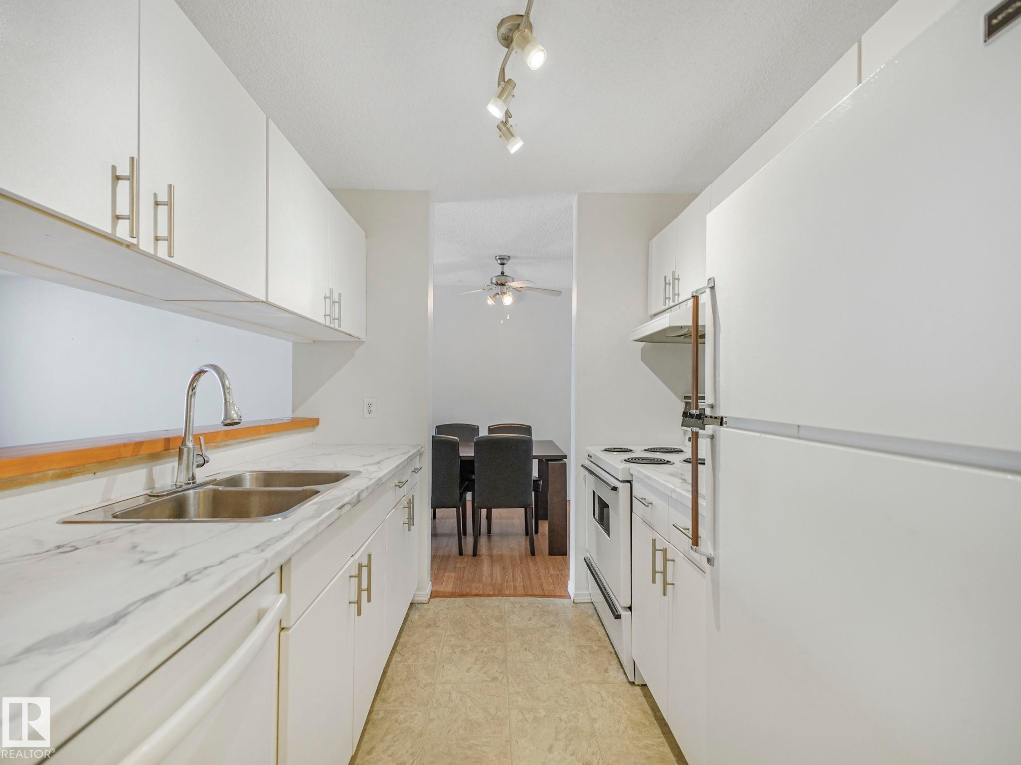 Galley kitchen featuring white cabinetry with silver-tone hardware, a double basin stainless steel sink, and light-toned countertop with veining - #143 10636 120 St Nw, Edmonton, AB - Indoor Photo Showing Kitchen With Double Sink